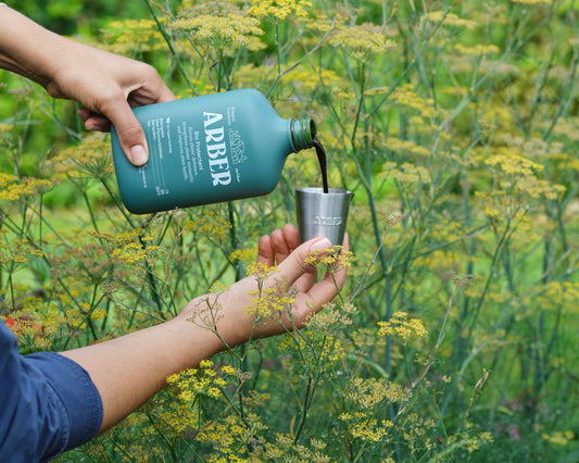 In a garden with tall yellow-green plants, an Arber Measuring Cup is used to pour liquid, highlighting this essential tool in action.