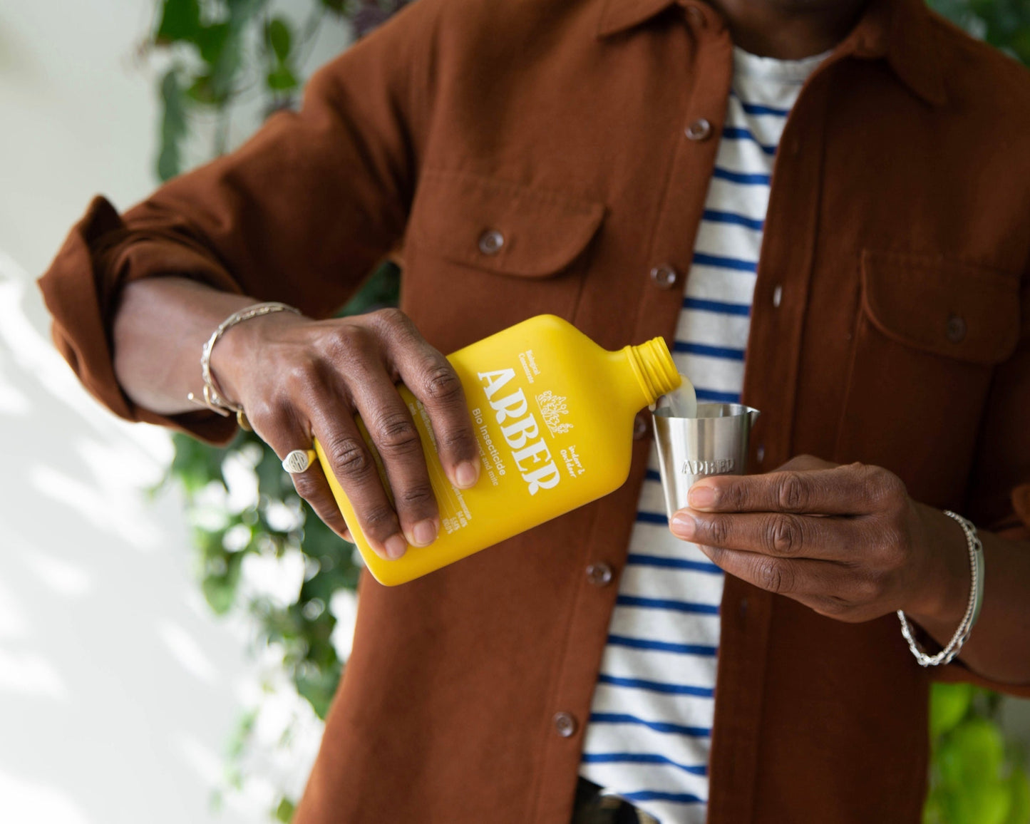Someone pours liquid from a yellow Arber bottle into a stainless steel Measuring Cup by Arber, surrounded by green foliage—a true garden essential.