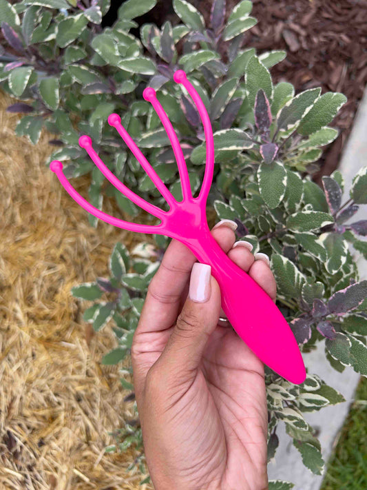 A hand with light pink nail polish holds the bright pink Memes Worms Worm Comb, a five-pronged scalp massager, in front of variegated leaves. This tool is designed to improve scalp aeration just like compost aerators.