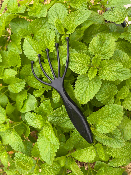 The Memes Worms Worm Comb, a small black plastic garden rake, rests on lush mint leaves—ideal for aerating compost and improving airflow in your garden beds.