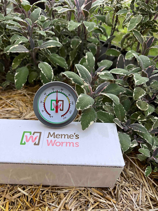 A Memes Worm Thermometer by Memes Worms reads about 75°F on a white box labeled Memes Worms, with straw and sage plants in the background for temperature monitoring in the worm farm.