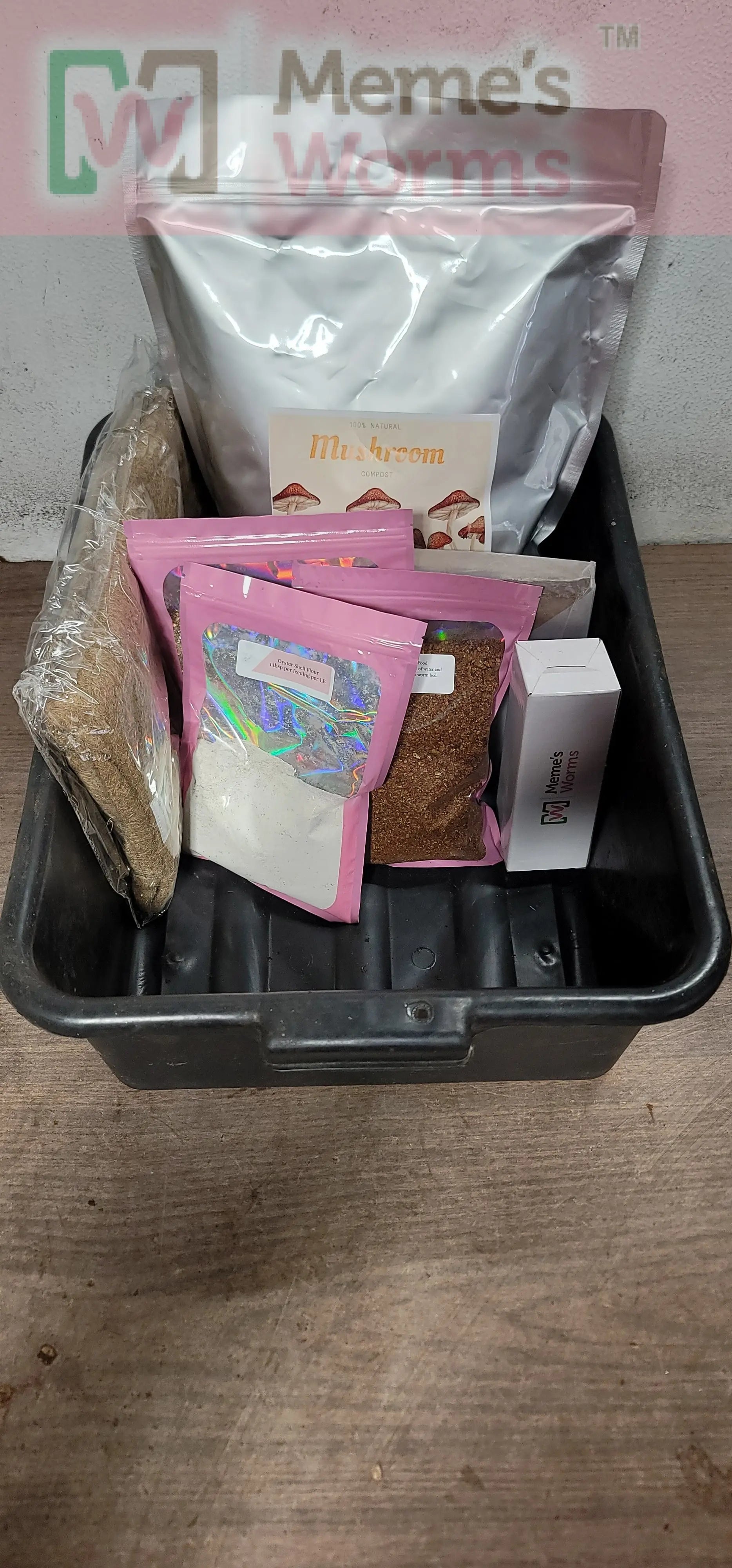The Worm Bin Starter Kit from Memes Worms is a black plastic bin filled with upright bags and packets of substrate, some labeled Mushroom. A partial sign reading Memes Worms appears behind the kit.