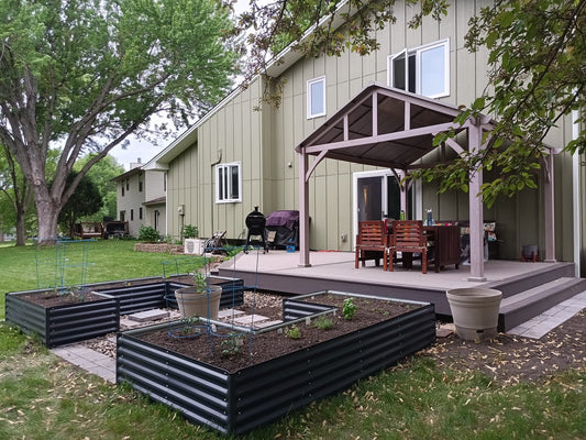 A backyard features an Ollegardens 17" Tall "U" Shape Infinity Raised Garden Bed, a patio with a wooden pergola, dining table, grill, and potted plants beside a light green house surrounded by trees and grass.