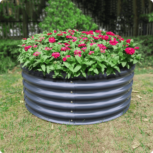 An Ollegardens 17" Tall x 42" Round Raised Garden Bed, filled with green plants and vibrant pink flowers, stands on a grassy lawn with trees and a fence in the background.