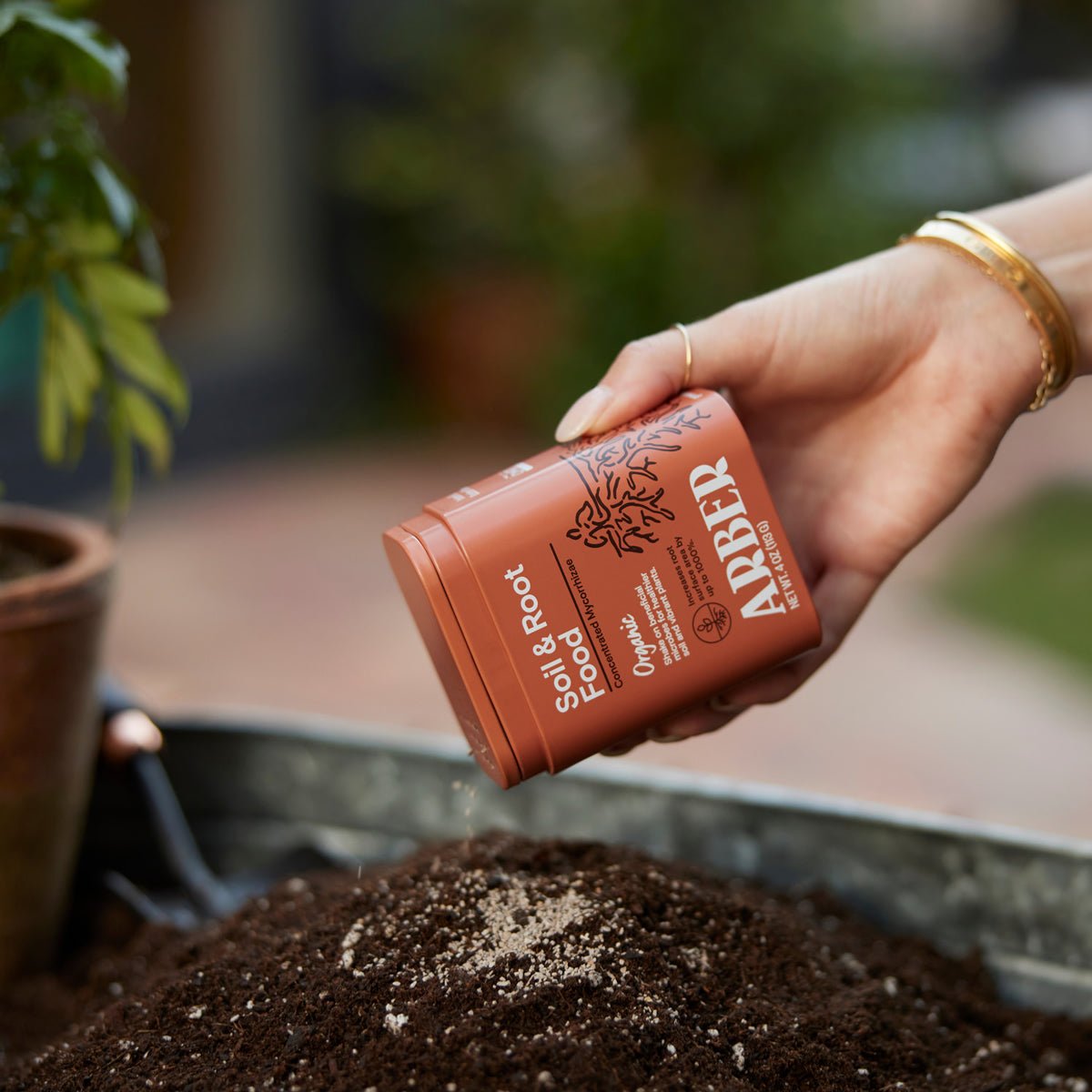 A hand sprinkles Arber Organic Soil & Root Boost, packed with beneficial microbes, onto potting soil in a garden scene, while a small plant in a terra cotta pot sits in the background.