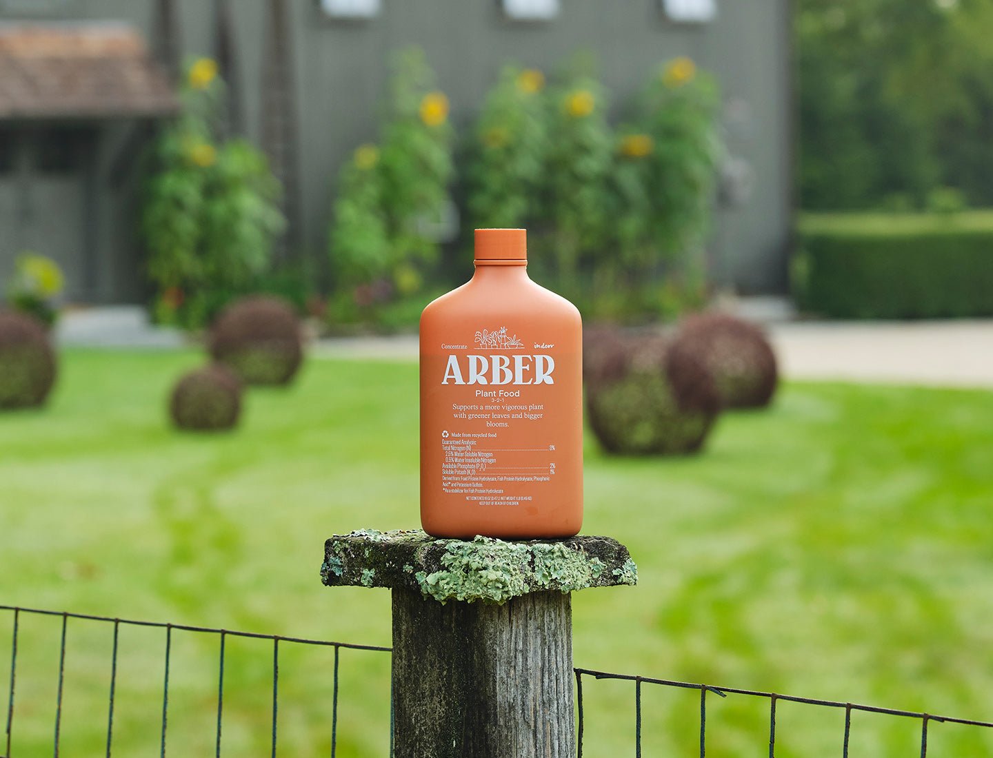 An orange bottle labeled Arber Organic Plant Food rests on a mossy wood post in a lush garden, emphasizing soil fertility with well-kept bushes and a softly blurred building in the distance.