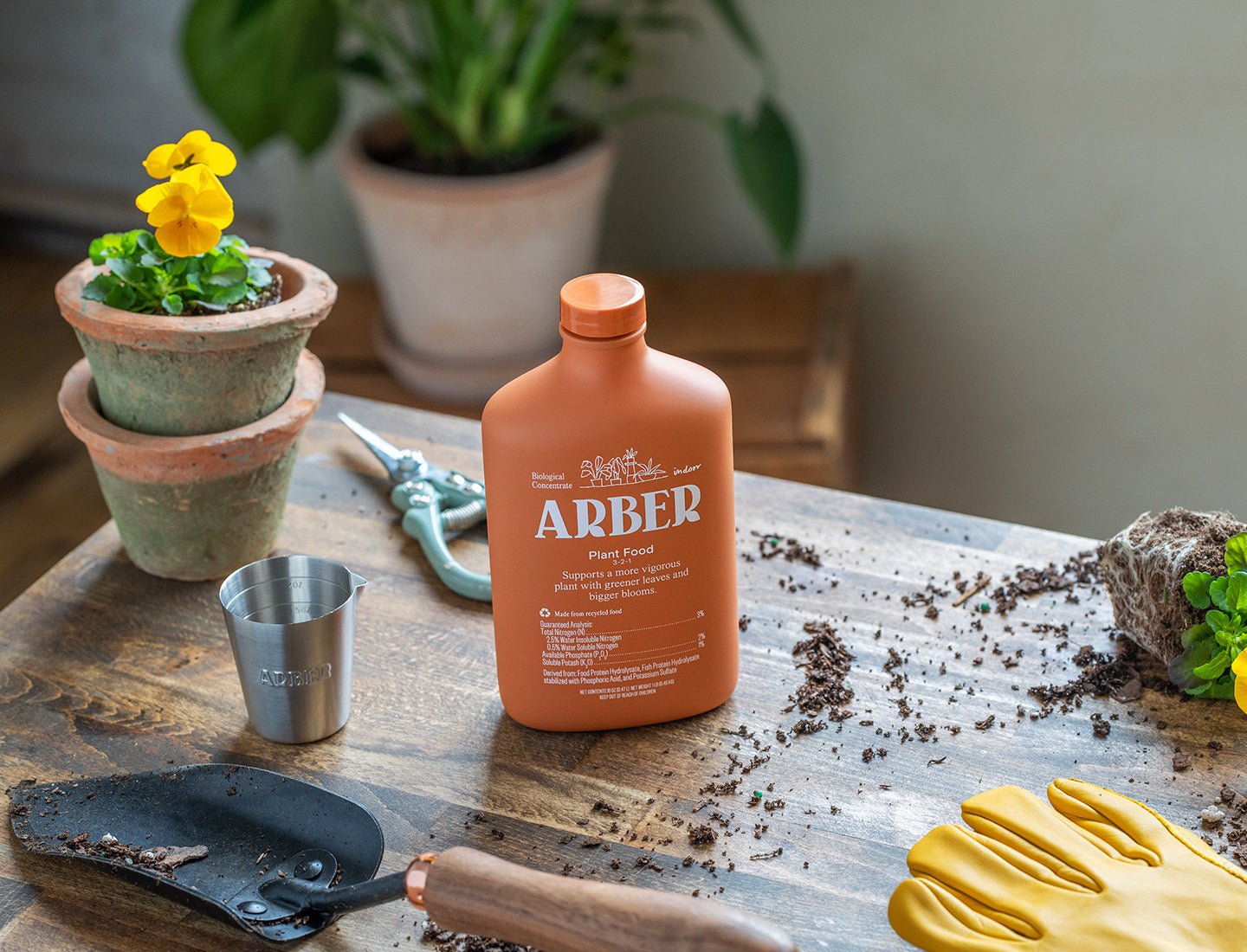 A bottle of Arber Organic Plant Food is placed on a wooden table with gardening tools, yellow gloves, a metal cup, potted flowers, and spilled soil—showcasing essential items for enriching soil during indoor gardening.