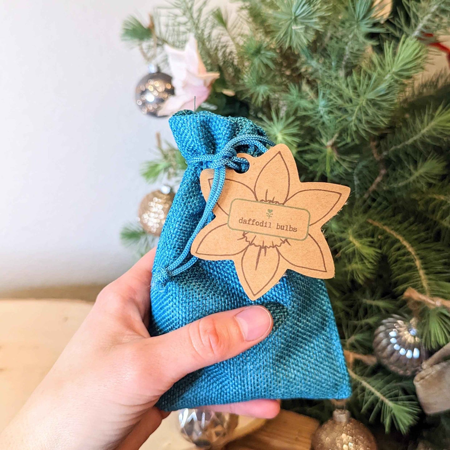 A hand holds a Plantflix Mini Daffodil Bulbs blue fabric bag with a daffodil-shaped tag, in front of a decorated Christmas tree—ideal for bringing cheerful spring blooms to your low-maintenance garden.