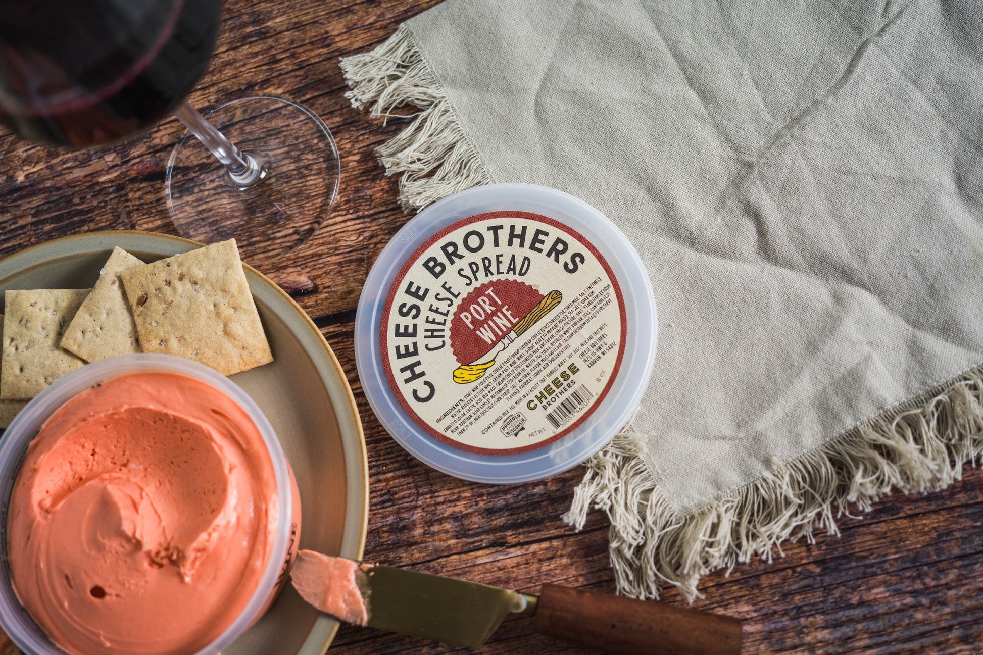 A container of Cheese Brothers, Inc. Port Wine Cheese Spread sits on a fringed cloth beside a plate with crackers, a knife, and cheese spread, with a glass of red wine on a wooden table.