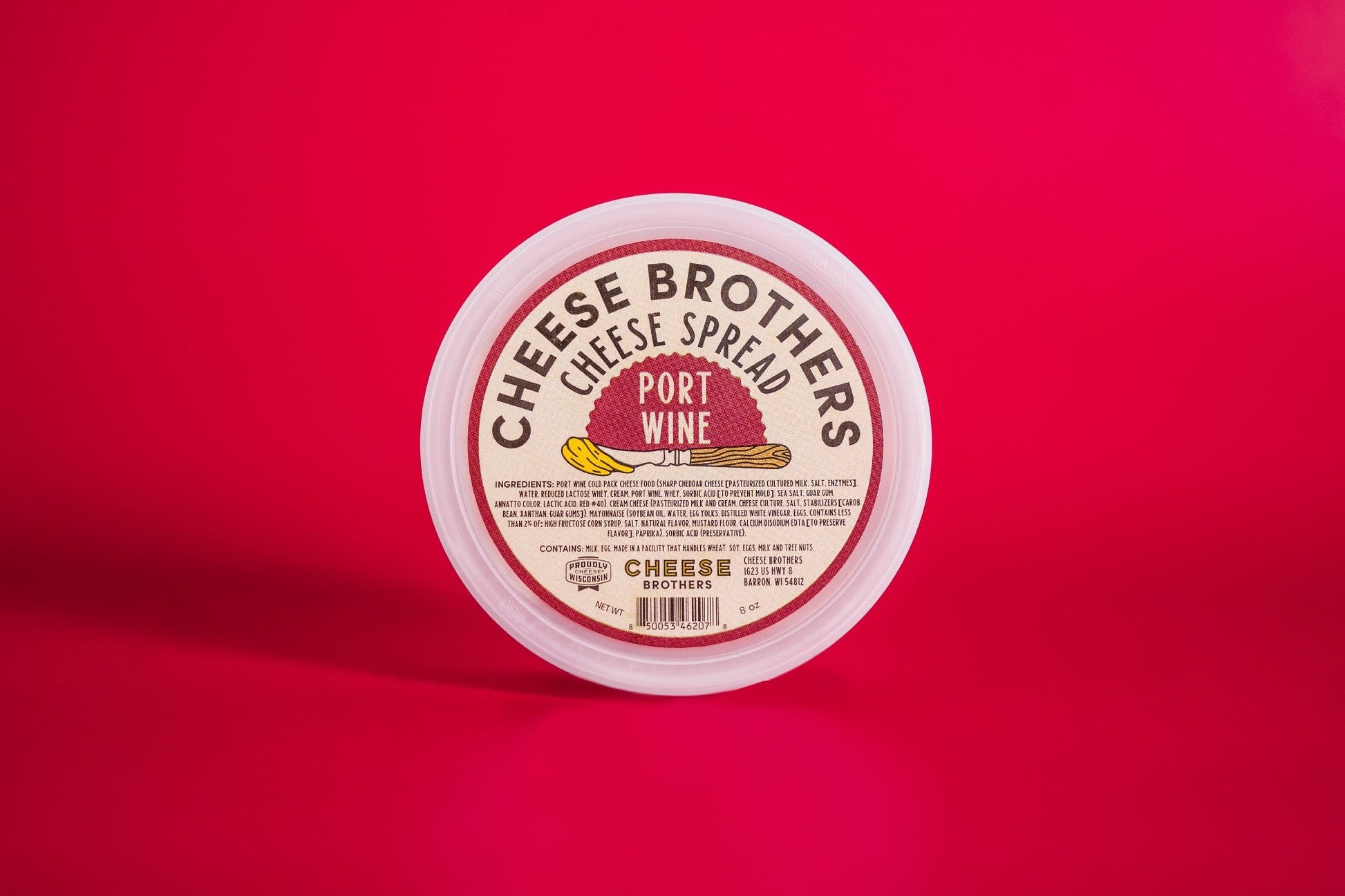 A round container of Cheese Brothers, Inc. Port Wine Cheese Spread sits on a bright red surface, its label showcasing an illustration of cheese and bold text highlighting creamy cheese made with Wisconsin cheddar.
