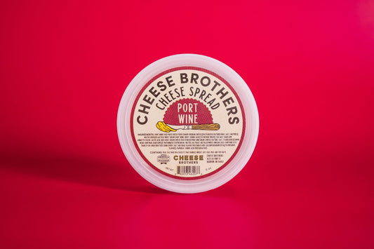 A round container of Cheese Brothers, Inc. Port Wine Cheese Spread sits on a bright red surface, its label showcasing an illustration of cheese and bold text highlighting creamy cheese made with Wisconsin cheddar.