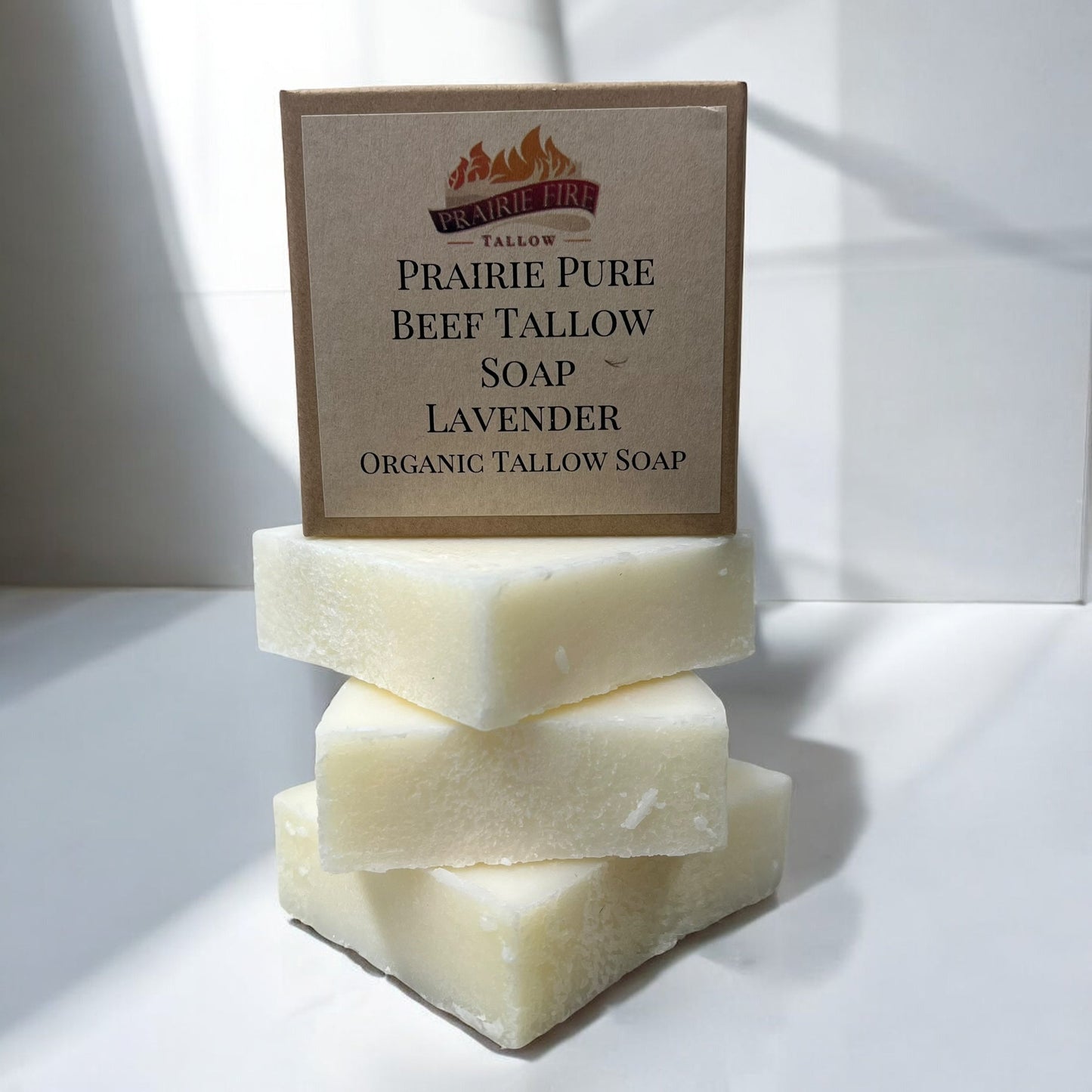 A Prairie Fire Tallow, Candles, and Lavender Pure Beef Tallow Soap Bar (3 Pack) box stands behind three stacked off-white, grass-fed tallow soap bars on a white surface with sunlight casting soft shadows. Natural, organic, USA-made.