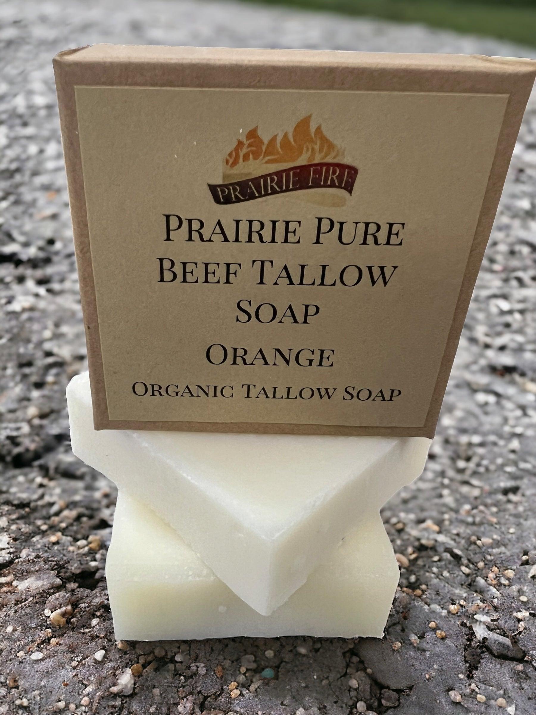 A box labeled Prairie Fire Tallow Pure Beef Tallow Soap Bar (3 Pack), made with grass-fed tallow in the USA, sits atop two white, natural and organic chemical-free soap bars on a rocky, textured outdoor surface.