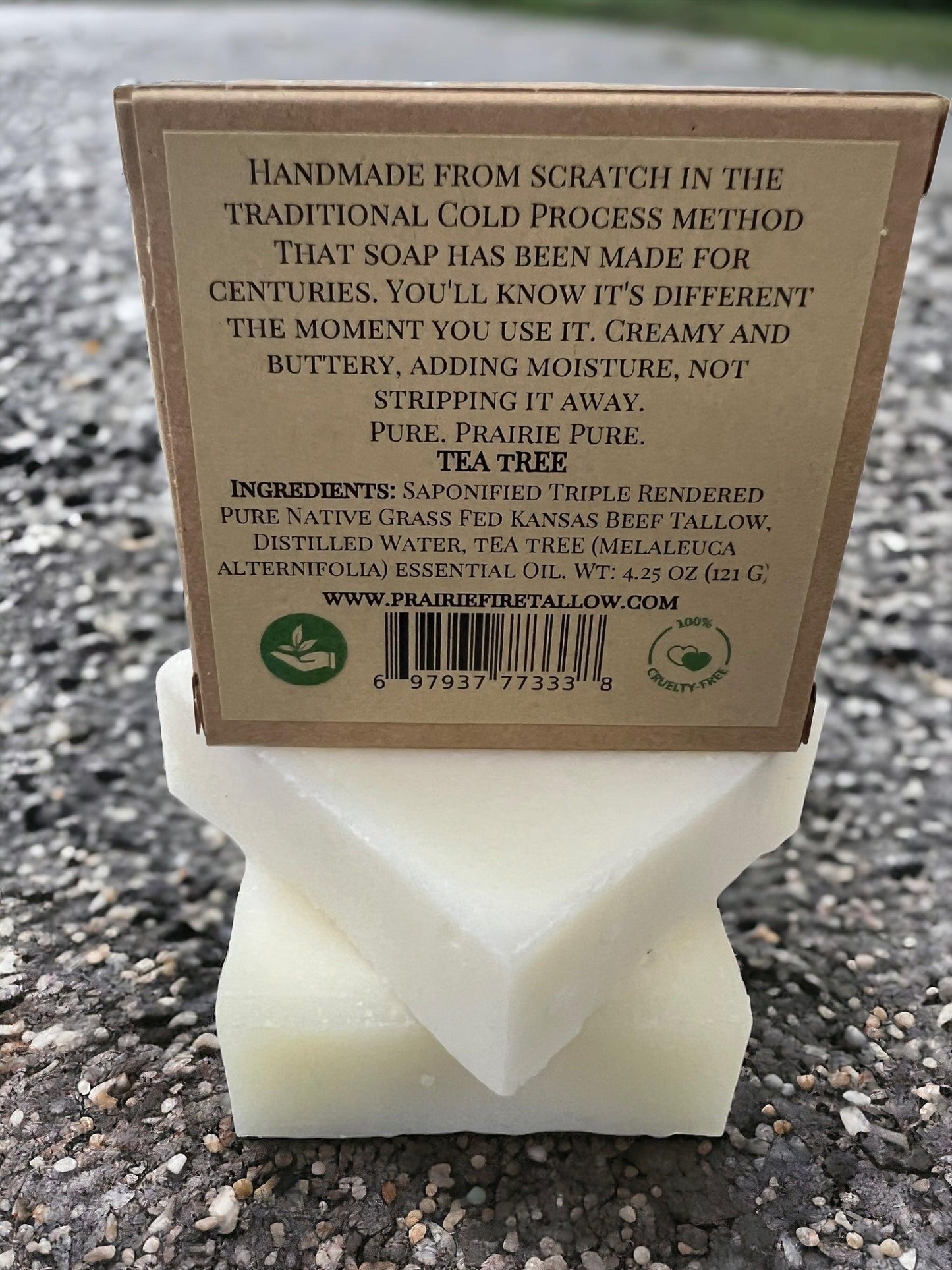 A Pure Beef Tallow Soap Bar from Prairie Fire Tallow, Candles, and Lavender rests on a textured surface, its label highlighting handmade cold process methods with beef tallow and tea tree oil. Ingredients and website are listed.