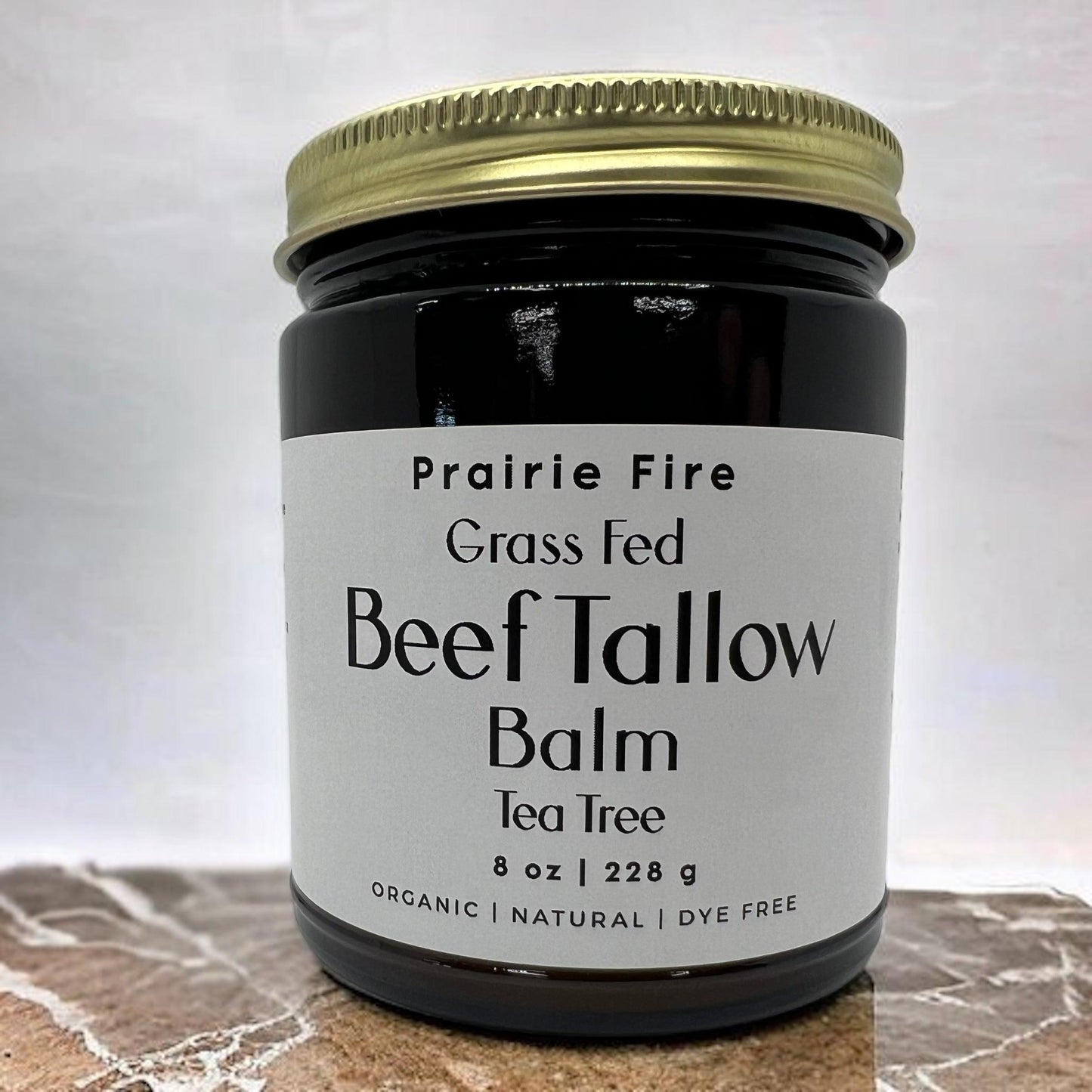 A dark glass jar with gold lid, labeled Prairie Fire Tallow, Candles, and Lavender Beef Tallow Balm - 8 oz, sits on a marble surface. This organic, grass-fed moisturizer is made in the USA for nourishing skin care.