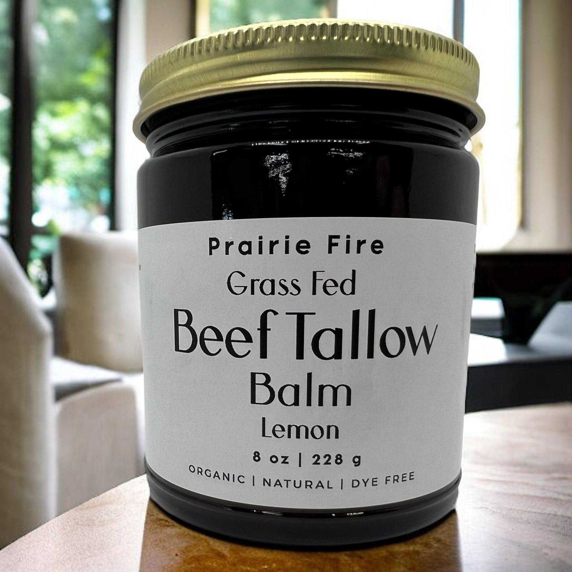 An 8 oz jar of Prairie Fire Tallow, Candles, and Lavender Beef Tallow Balm—organic, grass-fed, moisturizing skin care made in the USA—rests on a wooden surface with a modern blurred room in the background.