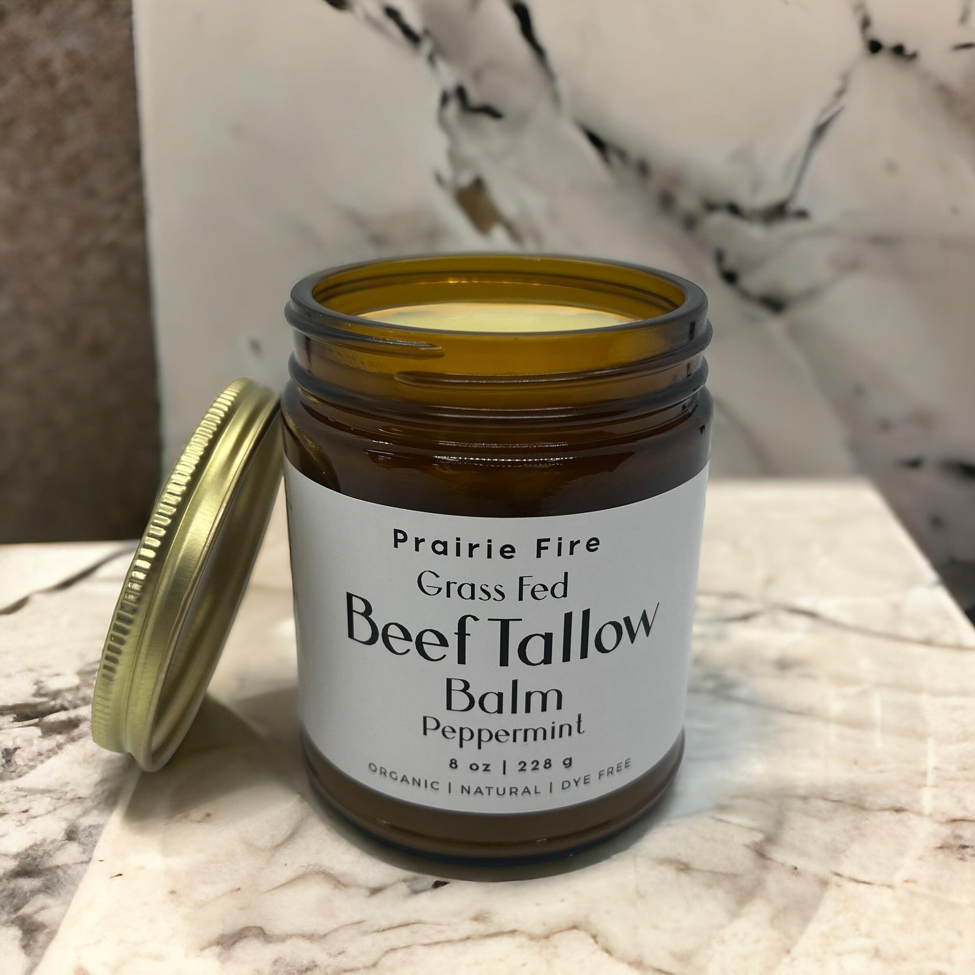 An 8 oz brown glass jar of Prairie Fire Tallow, Candles, and Lavender’s Organic Grass Fed Beef Tallow Balm sits on marble with its lid nearby. The label notes this USA-made balm as a natural, dye-free moisturizer for skin care.