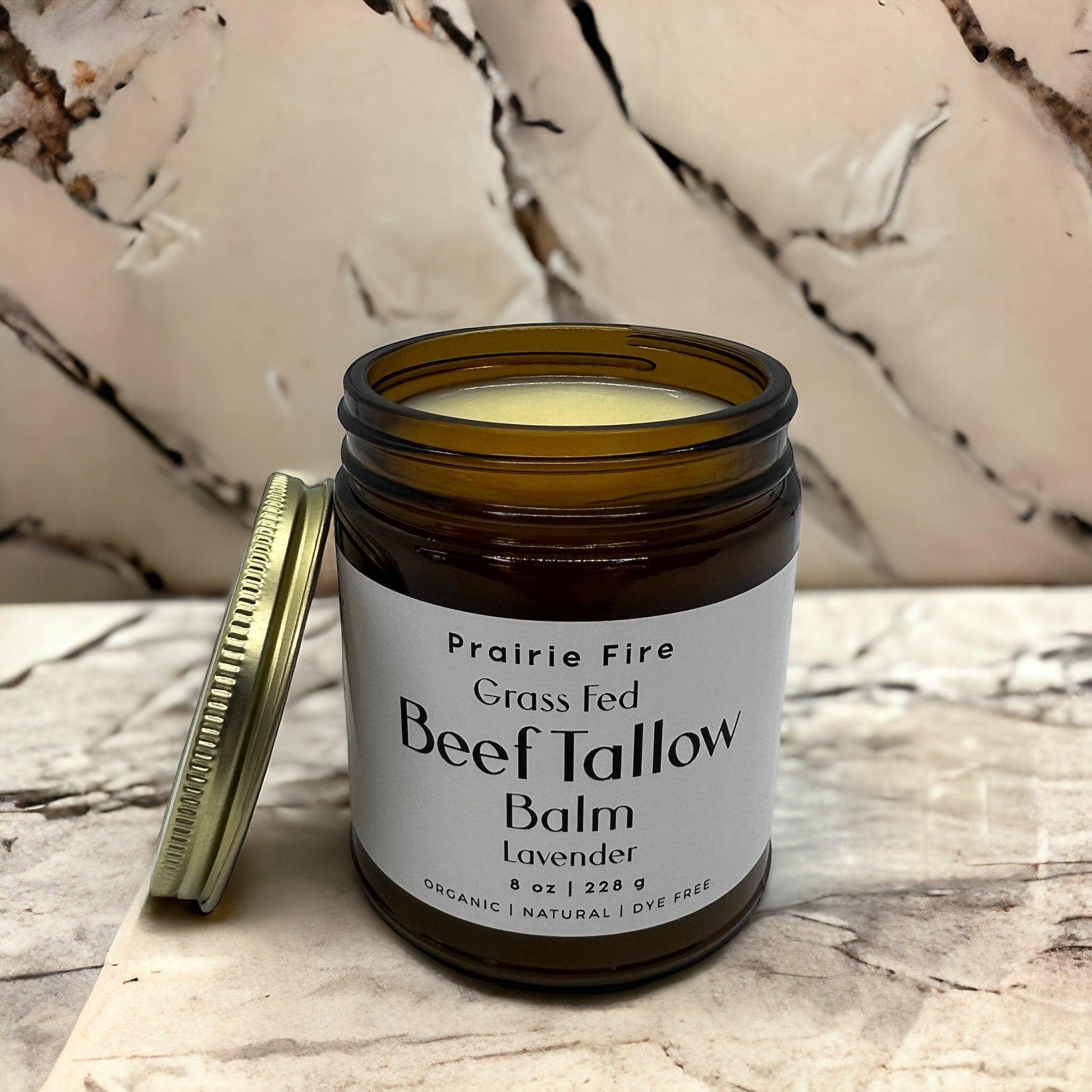 An open 8 oz brown glass jar of Prairie Fire Tallow, Candles, and Lavender Beef Tallow Balm sits with its lid beside it on marble. This organic, grass fed and finished USA-made moisturizer features smooth light yellow balm inside.