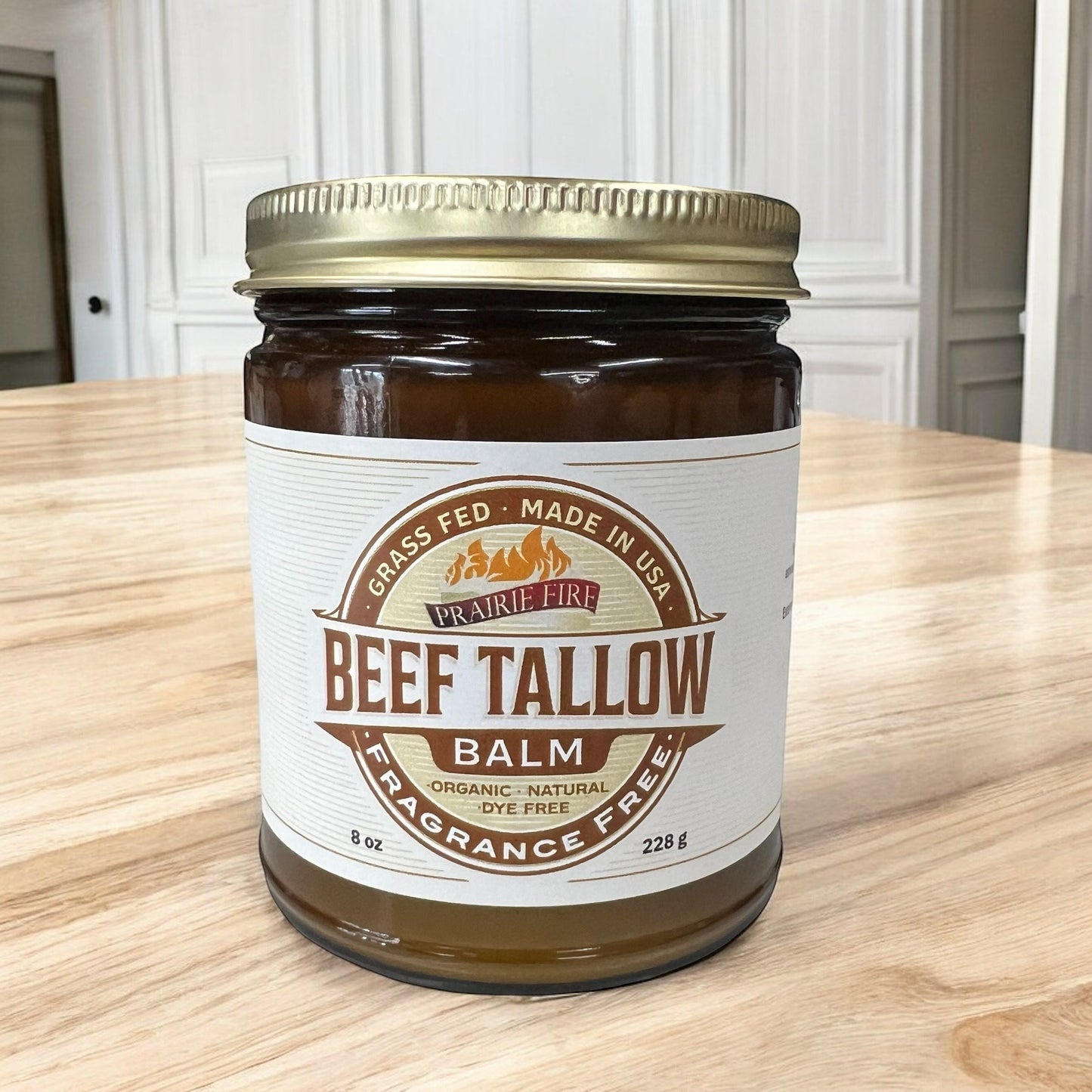 An 8 oz jar of Prairie Fire Tallow, Candles, and Lavender Beef Tallow Balm with a gold lid sits on a wooden table. This organic, grass-fed moisturizer is dye- and fragrance-free, and proudly made in the USA.