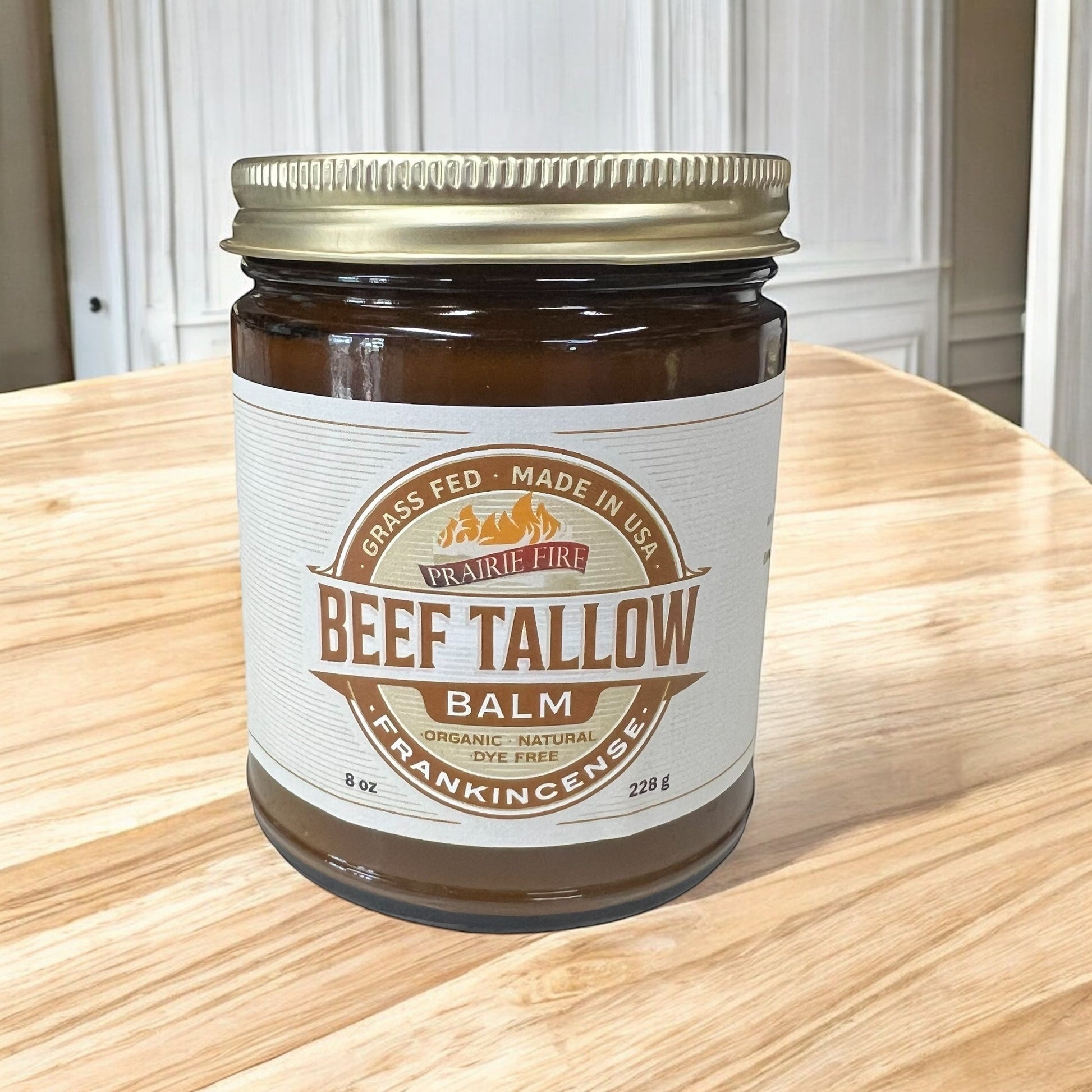 An 8 oz jar of Prairie Fire Tallow Beef Tallow Balm with a gold lid rests on wood. The organic, dye-free moisturizer is made in the USA from grass-fed, finished tallow and frankincense by Prairie Fire Tallow, Candles, and Lavender.