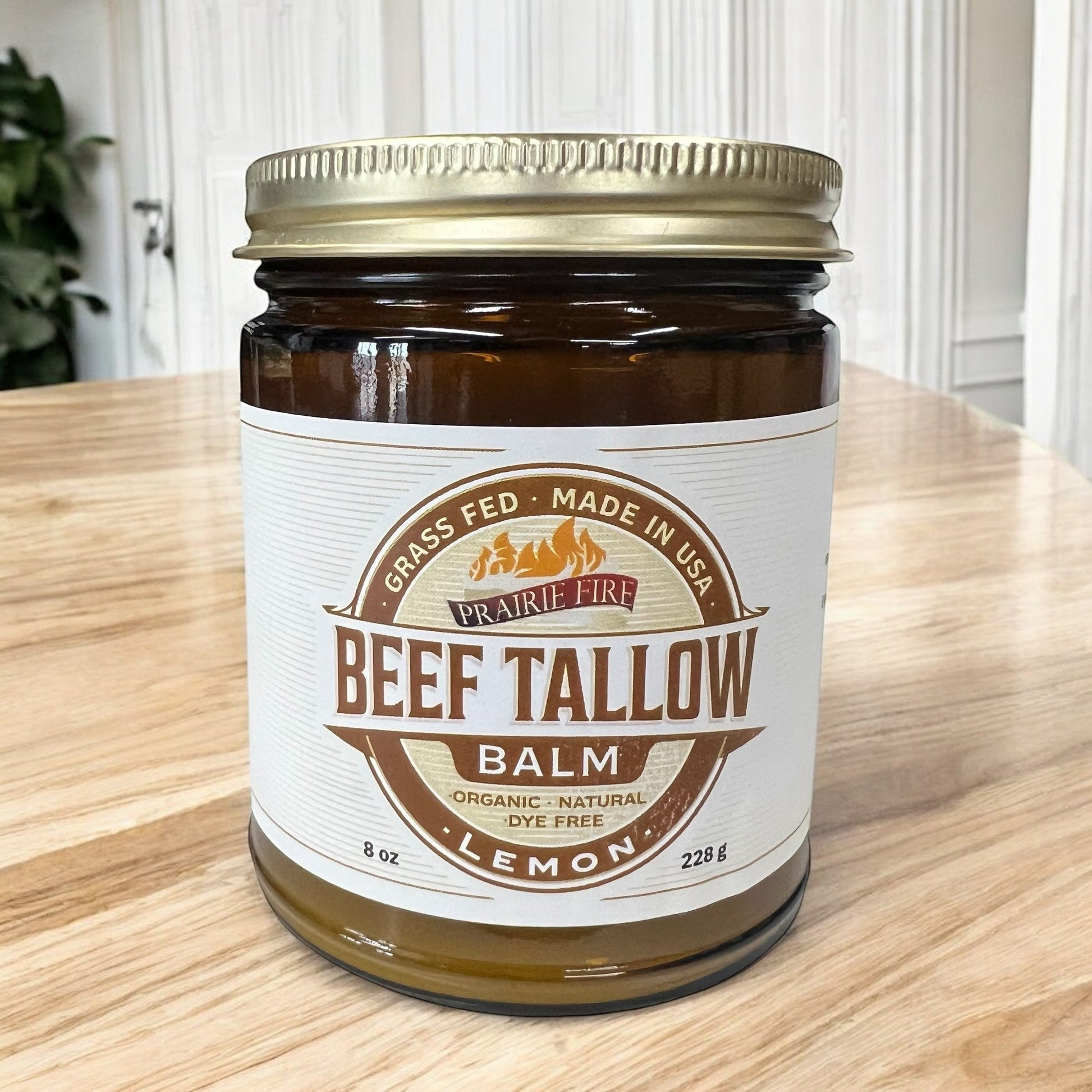An 8 oz glass jar of Prairie Fire Tallow, Candles, and Lavender Beef Tallow Balm sits on a wooden surface. This organic, grass-fed moisturizer is made in the USA, lemon-scented, dye-free, and topped with a gold lid.