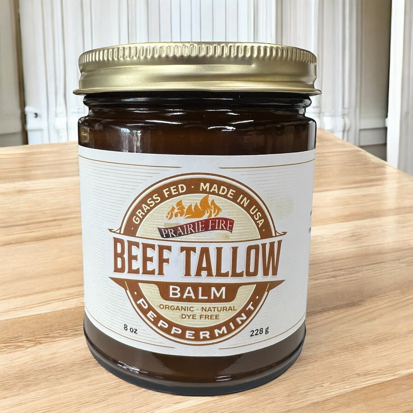A brown glass jar labeled Prairie Fire Tallow, Candles, and Lavender Beef Tallow Balm - 8 oz sits on wood. This organic, grass-fed and finished moisturizer is dye-free, made in the USA, and ideal for natural skin care.