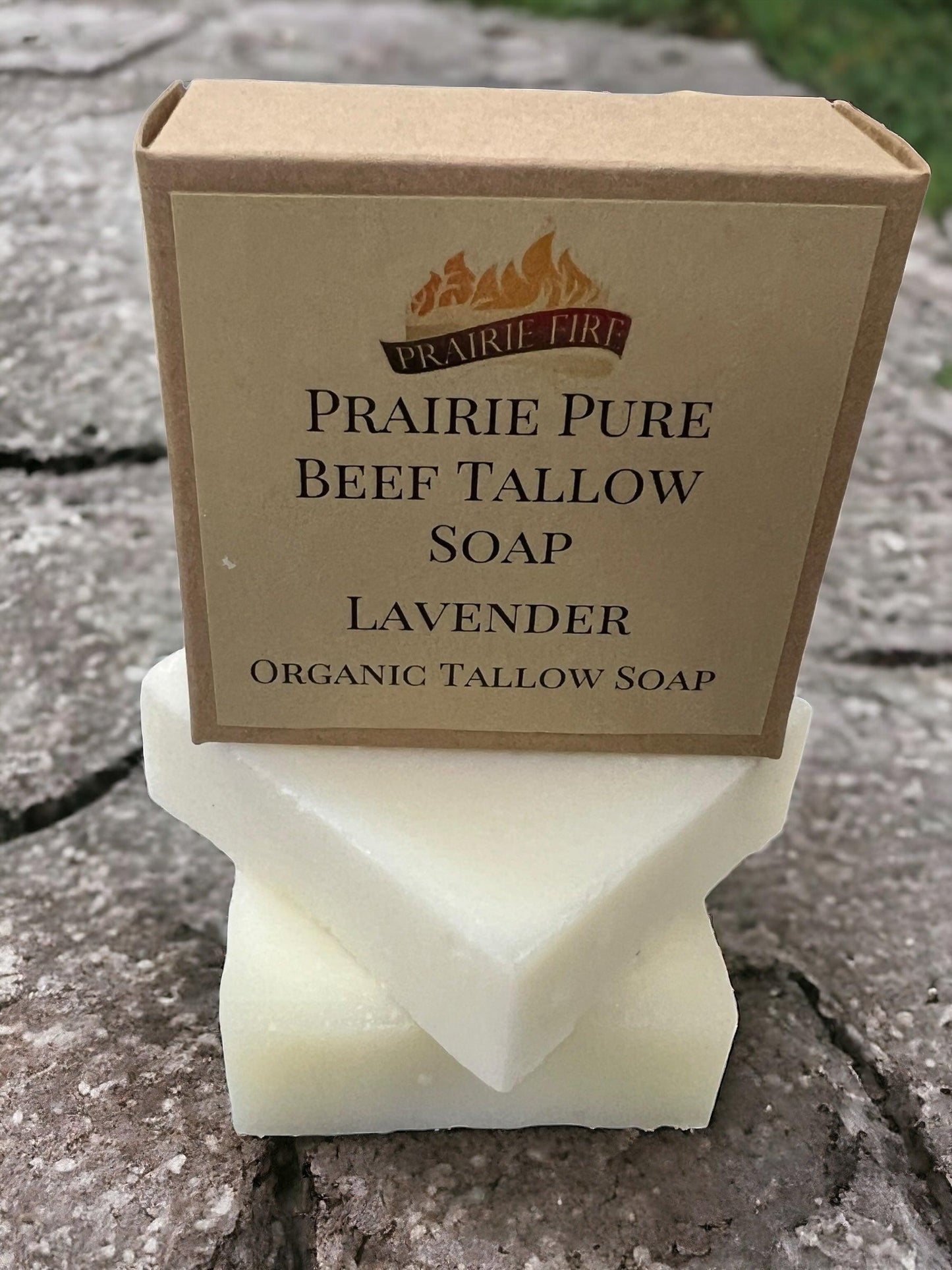 A Prairie Fire Tallow Pure Beef Tallow Soap Bar (3 Pack), Grass Fed and Finished, rests on two white bars outdoors. The brown packaging with clear labeling and a flame logo highlights its natural, organic ingredients for face, body, and hair.