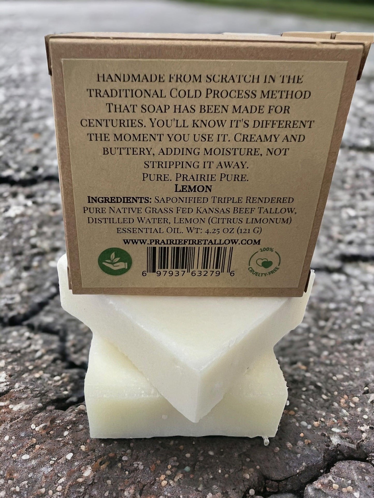 A Pure Beef Tallow Soap Bar by Prairie Fire Tallow, Candles, and Lavender with a brown label sits on cracked pavement. This natural, organic soap is handmade in the USA with grass-fed tallow and essential oils for face, body, and hair.