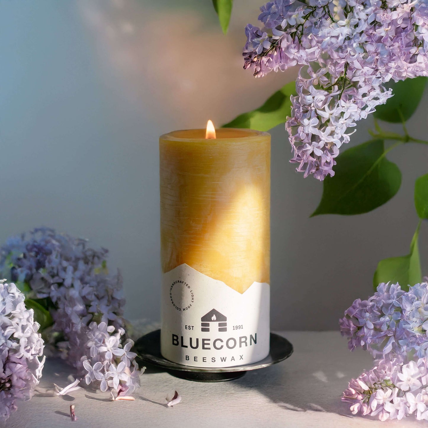 Bluecorn Candles pure beeswax pillar candle held in a handmade holder during golden hour with spring lilacs in the background.