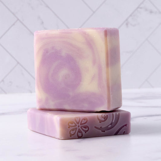 Two square Rustic Strength Bar Soap | Lavender bars are stacked on marble; the front, perfect for sensitive skin, displays engraved floral and swirl designs. A white tiled wall forms the background.