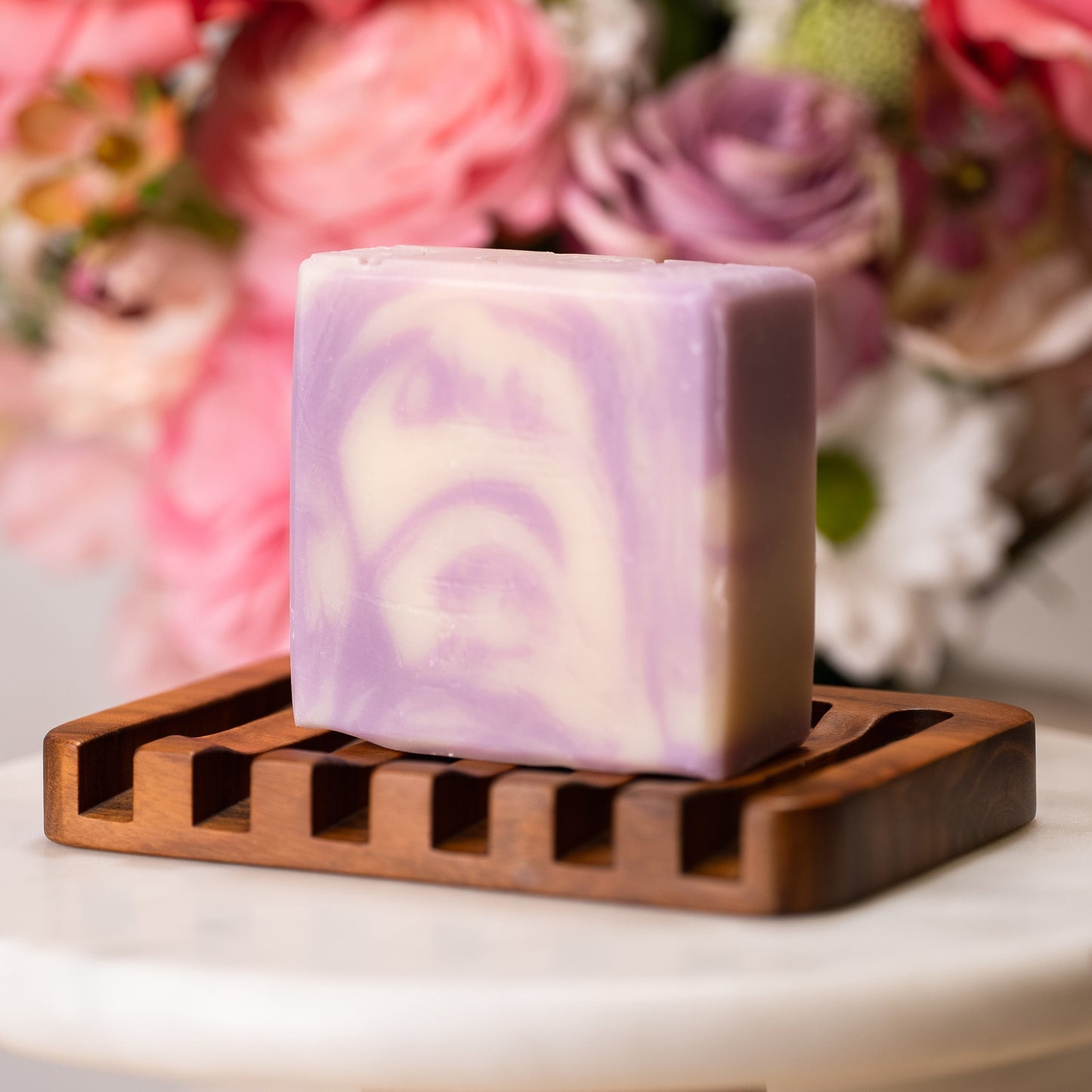 Rustic Strength’s Bar Soap | Lavender—a square, purple and white marbled bar—rests on a wooden soap dish with soft-focus pink and white flowers behind. Perfect for sensitive skin.