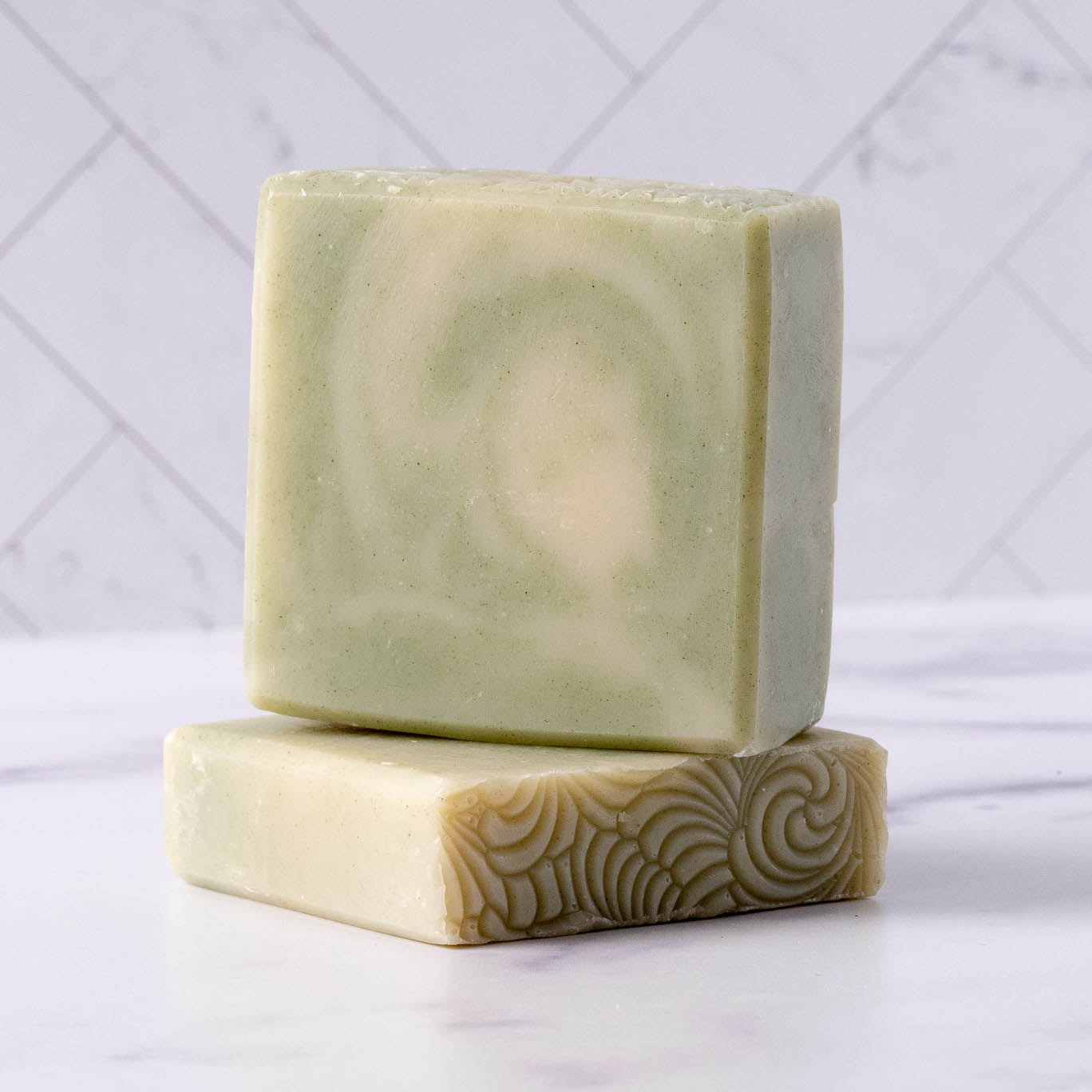 Two greenish-beige Rustic Strength Bar Soap | Peppermint Tea Tree bars sit on a white surface, one upright with a subtle swirl and the other flat showing texture, set against a marble tile background.
