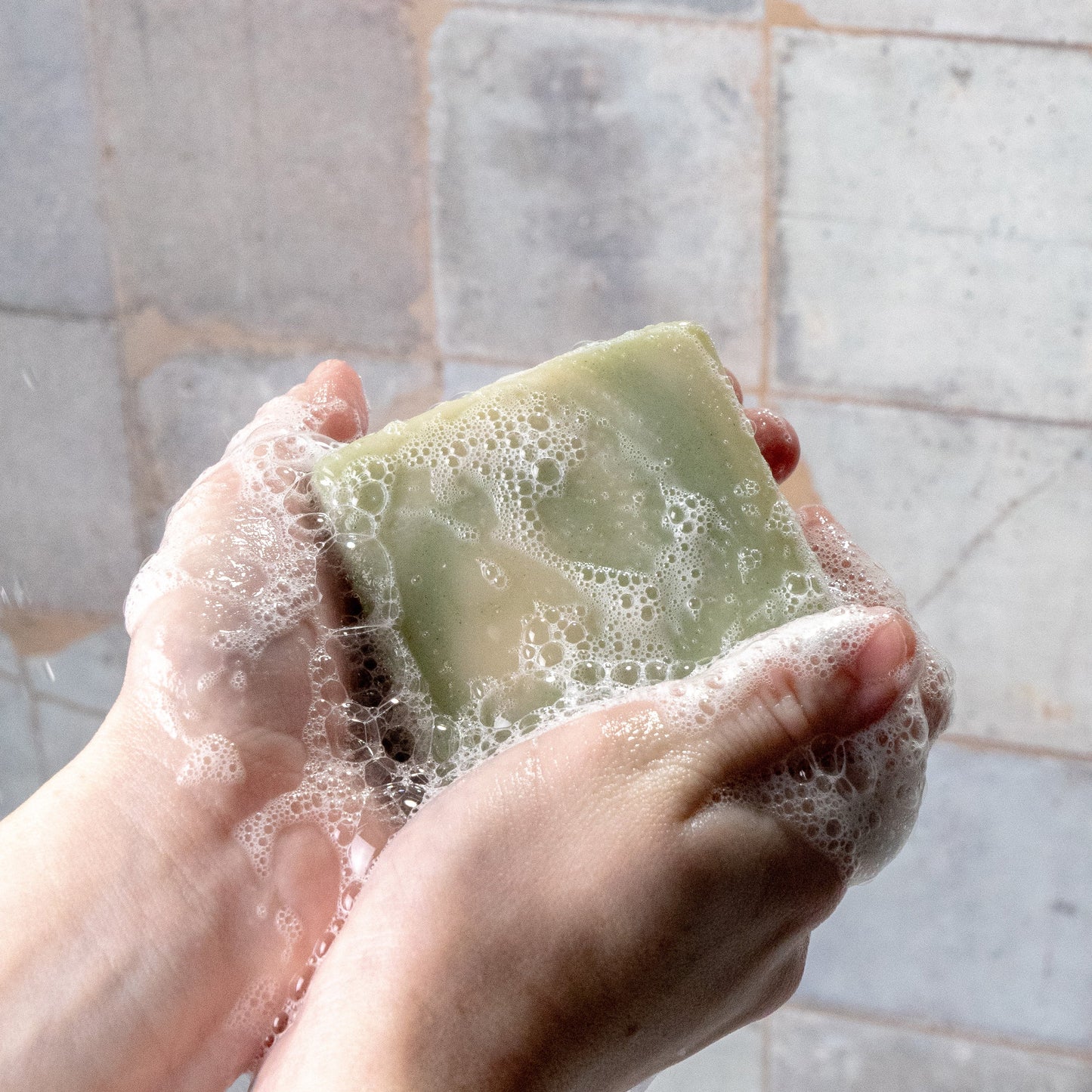 A person holds Rustic Strengths Bar Soap | Peppermint Tea Tree in both hands, lathering it into white, foamy bubbles. Pale, textured tiles appear in the background.