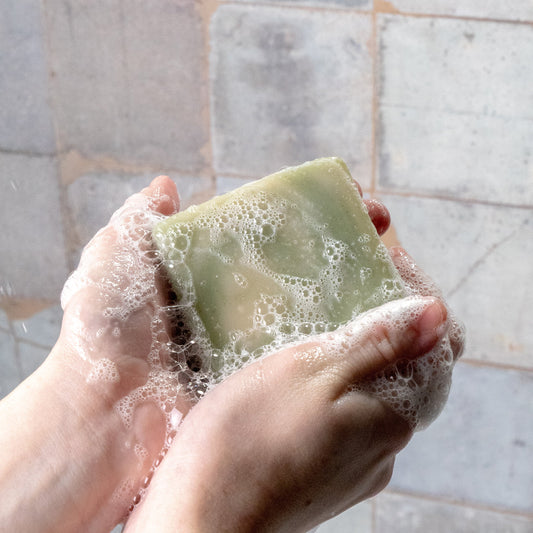A person holds Rustic Strengths Bar Soap | Peppermint Tea Tree in both hands, lathering it into white, foamy bubbles. Pale, textured tiles appear in the background.