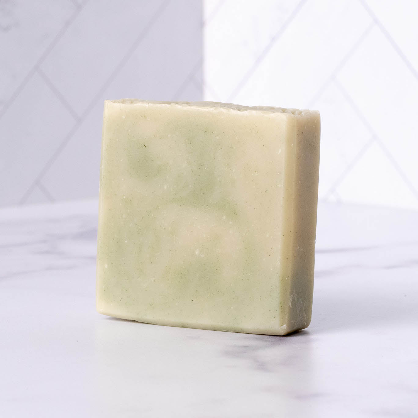 A rectangular bar of Rustic Strength Bar Soap | Peppermint Tea Tree in pale green with subtle swirls stands upright on a white marble surface, set against a white tiled background.