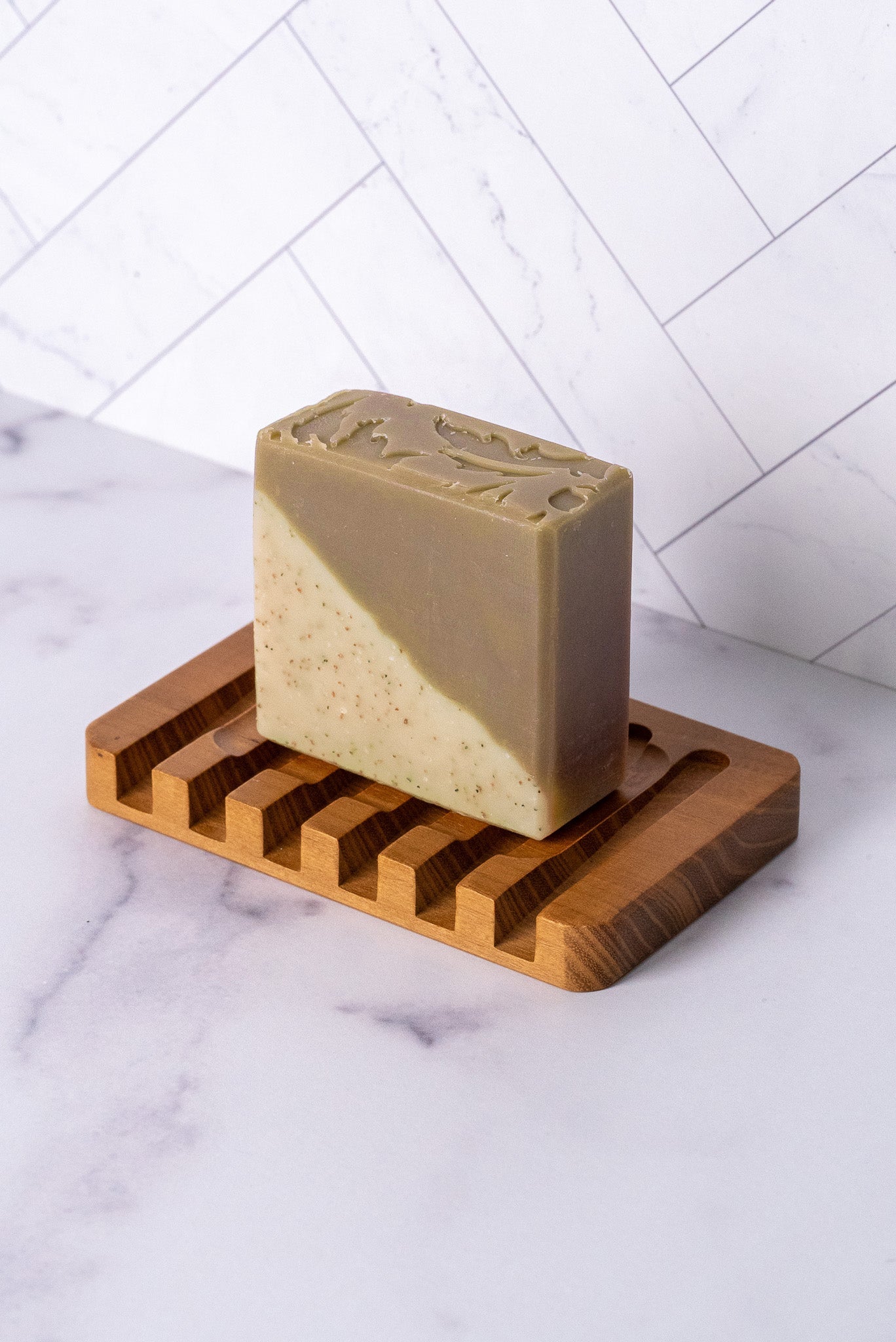 The Rustic Strength Bar Soap | Sandalwood Patchouli, a rectangular cold process bar with two-tone diagonal colors and hydrating natural ingredients, rests on a grooved wooden soap dish atop marble before a white tile wall.