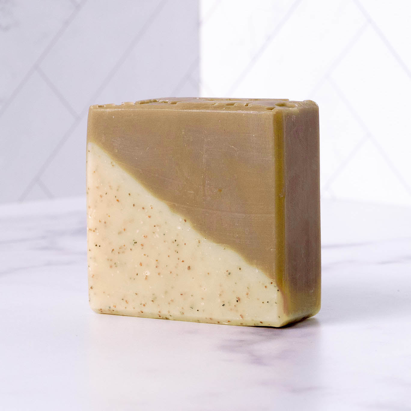 The Rustic Strength Bar Soap | Sandalwood Patchouli is a rectangular, two-toned cold process bar standing upright on a white, marble-like surface. One diagonal half is beige; the other, light brown with exfoliating walnut speckles.