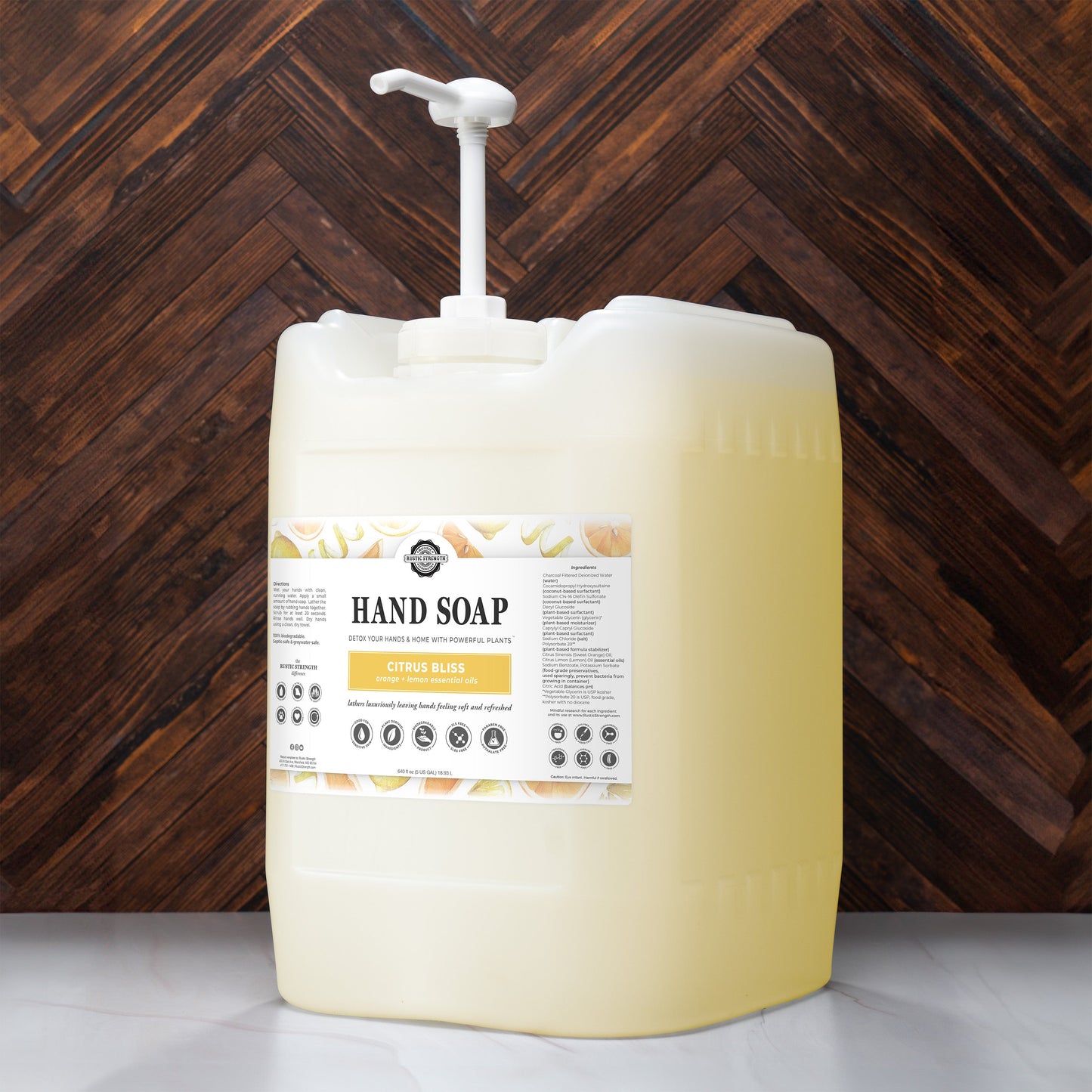 A large plastic pump bottle of Rustic Strength Hand Soap | Citrus Bliss sits on a surface before a dark chevron-patterned wood wall. The label reads Hand Soap | Citrus Bliss and displays images of citrus fruits.