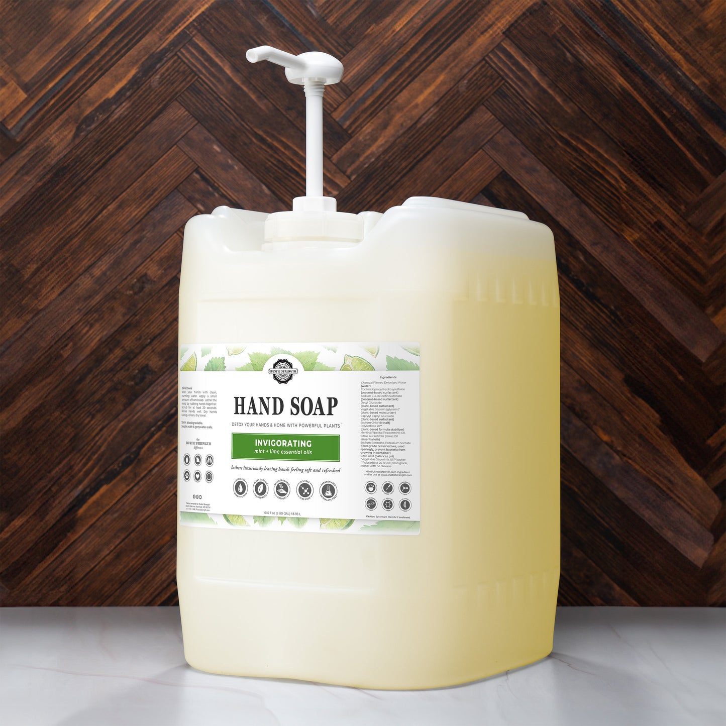 A large, translucent plastic pump bottle of Rustic Strength Hand Soap | Invigorating with yellow biodegradable liquid and green leaf graphics on the label sits on a white surface against a dark wood background.