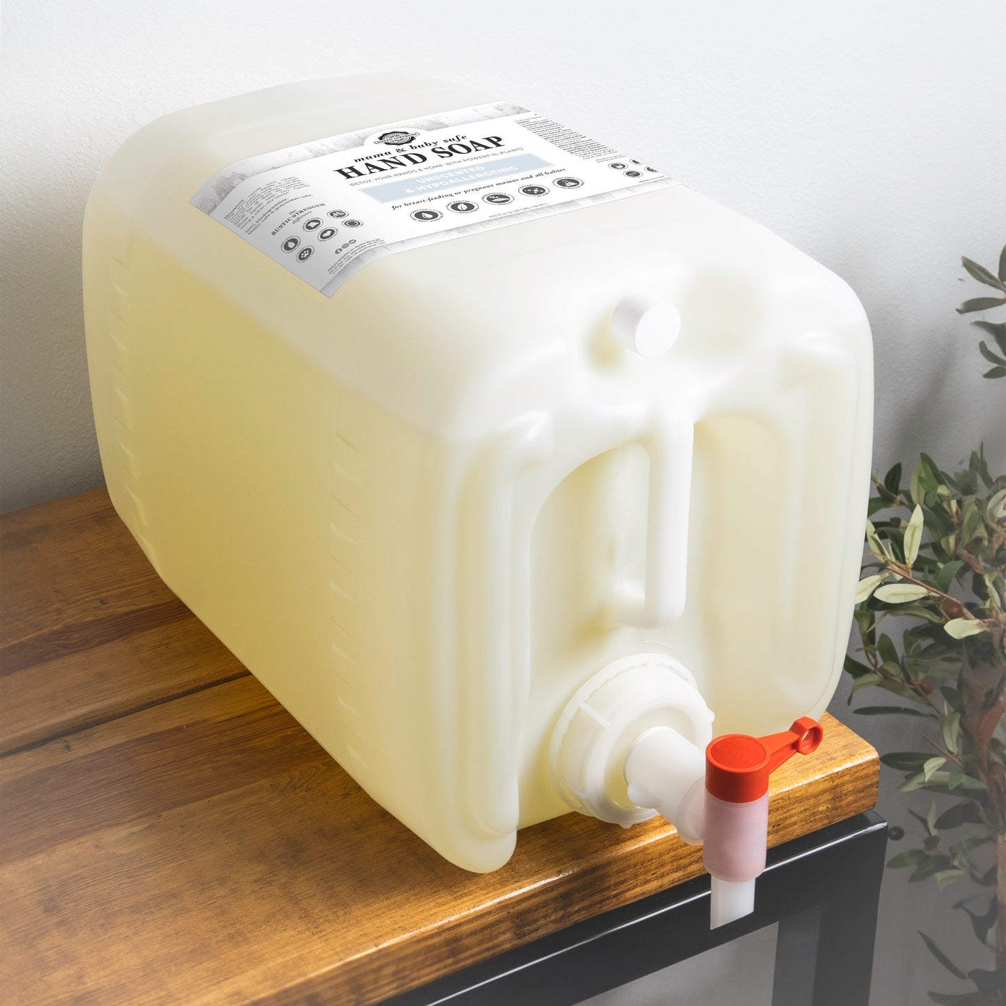 A large rectangular Rustic Strength Hand Soap | Mama + Baby Safe container with a white label and red/white spigot sits on a wooden surface near a plant, ready for dispensing.