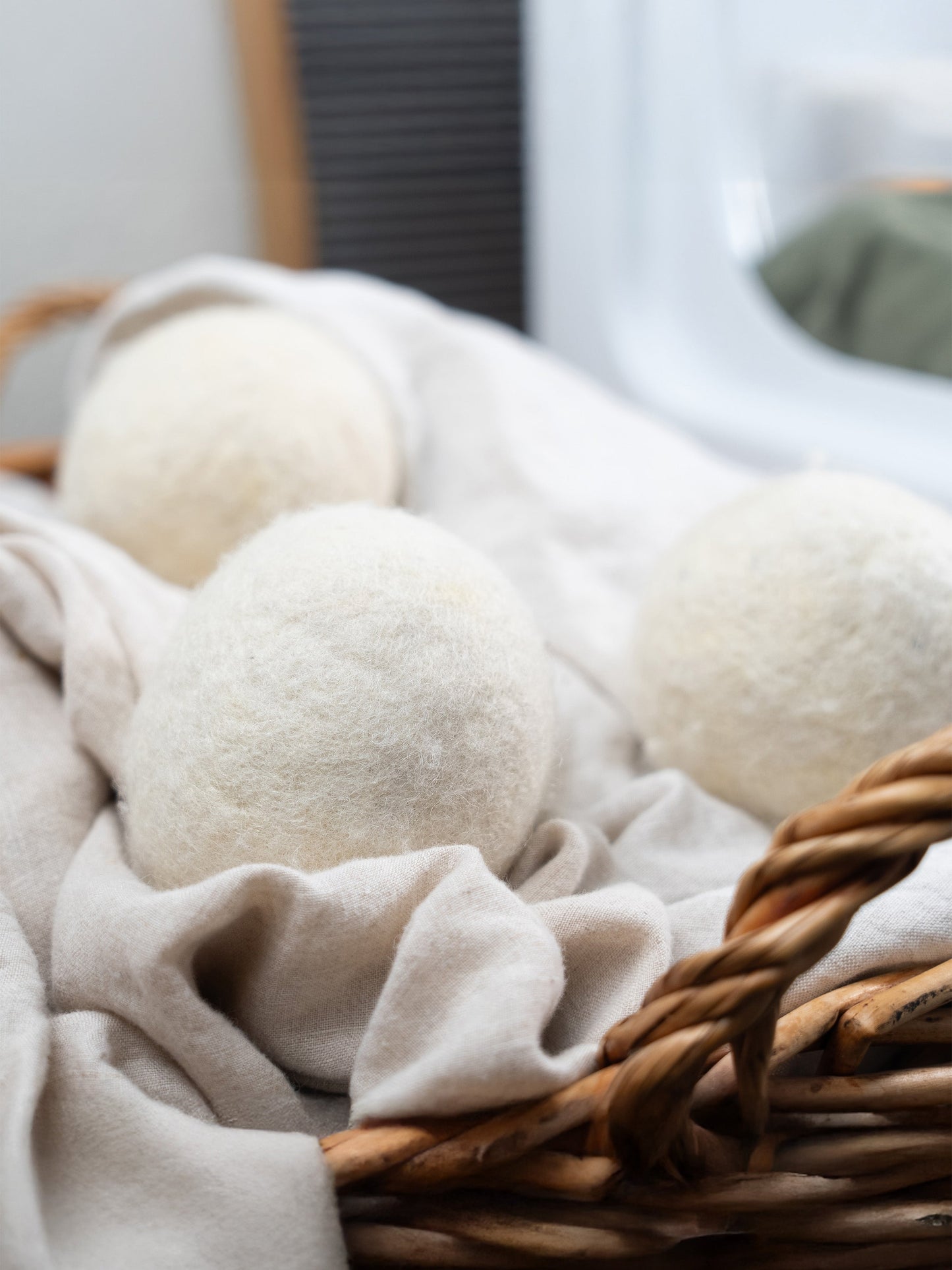 Three Jumbo Wool Dryer Balls (3 pack) from Rustic Strength sit on a beige cloth in a basket, with laundry and a washer in the background. Made using regenerative agriculture, these eco-friendly balls provide a sustainable laundry solution.