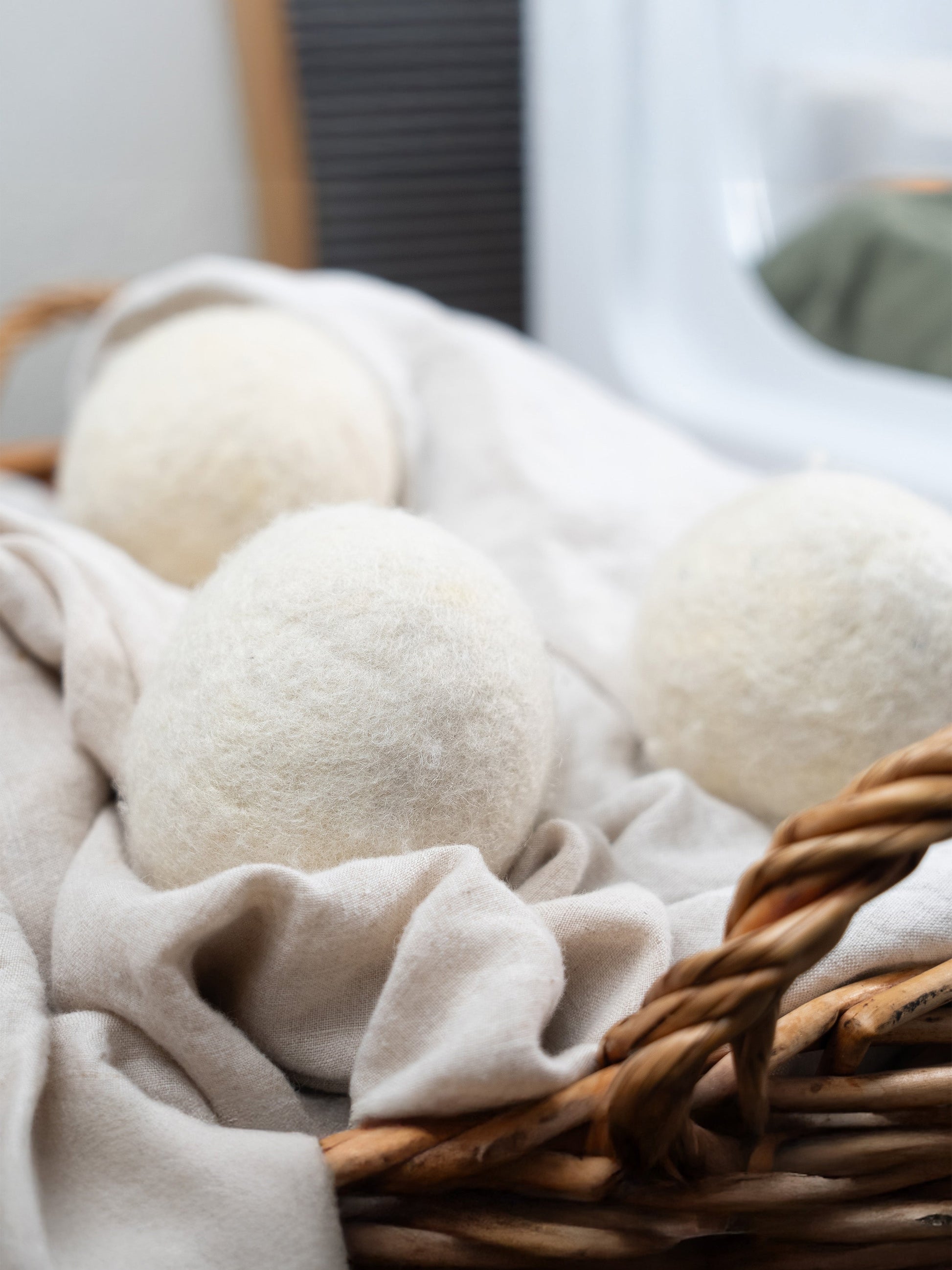 Three Jumbo Wool Dryer Balls (3 pack) from Rustic Strength sit on a beige cloth in a basket, with laundry and a washer in the background. Made using regenerative agriculture, these eco-friendly balls provide a sustainable laundry solution.