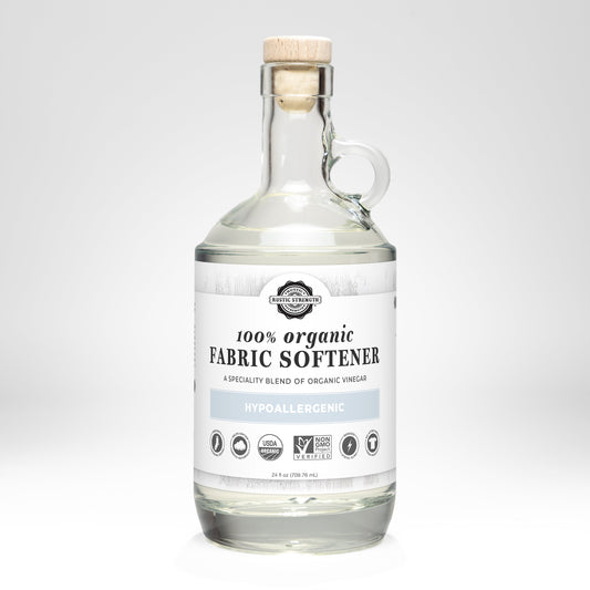 Rustic Strength Organic Fabric Softener comes in a clear glass bottle with a cork stopper and white label featuring hypoallergenic, biodegradable, plant-based ingredients, certification symbols, and a soft blue accent.