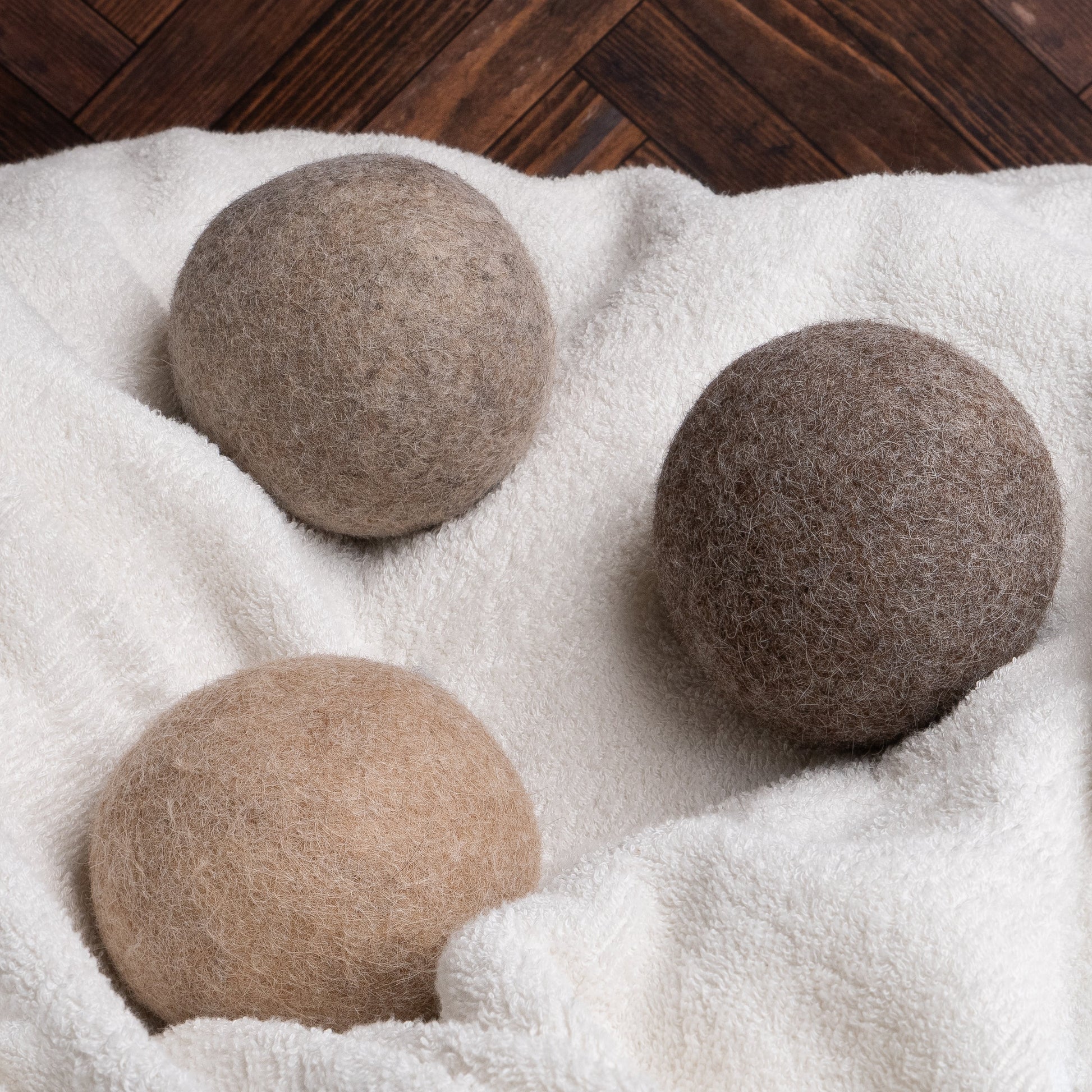 Rustic Strength Alpaca Dryer Balls (3 pack) in assorted brown shades rest on soft, white fabric with a dark wooden floor in the background, providing natural static reduction for your laundry.