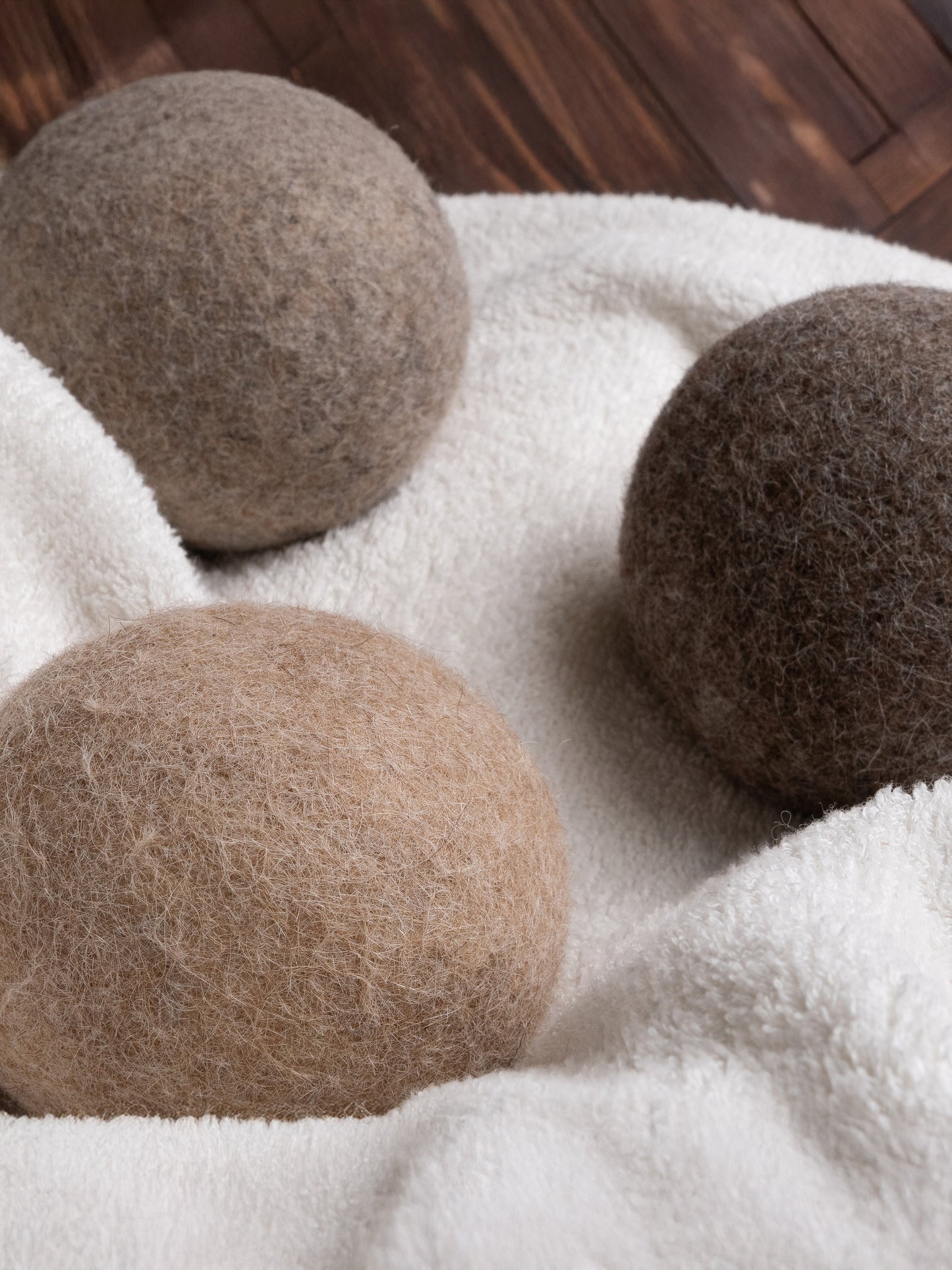 Rustic Strength Alpaca Dryer Balls (3 pack) in brown and gray rest on soft white fabric, providing a natural wool alternative for laundry and reducing static. A wooden floor is visible in the background.