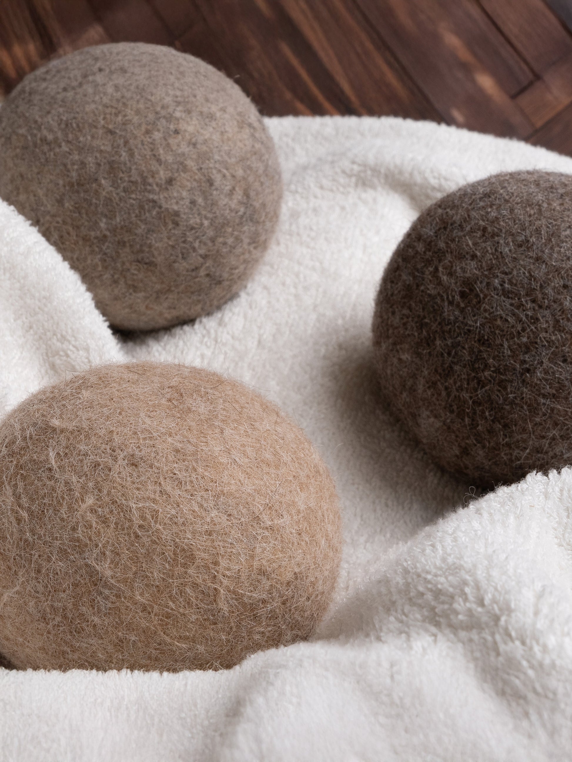 Rustic Strength Alpaca Dryer Balls (3 pack) in brown and gray rest on soft white fabric, providing a natural wool alternative for laundry and reducing static. A wooden floor is visible in the background.