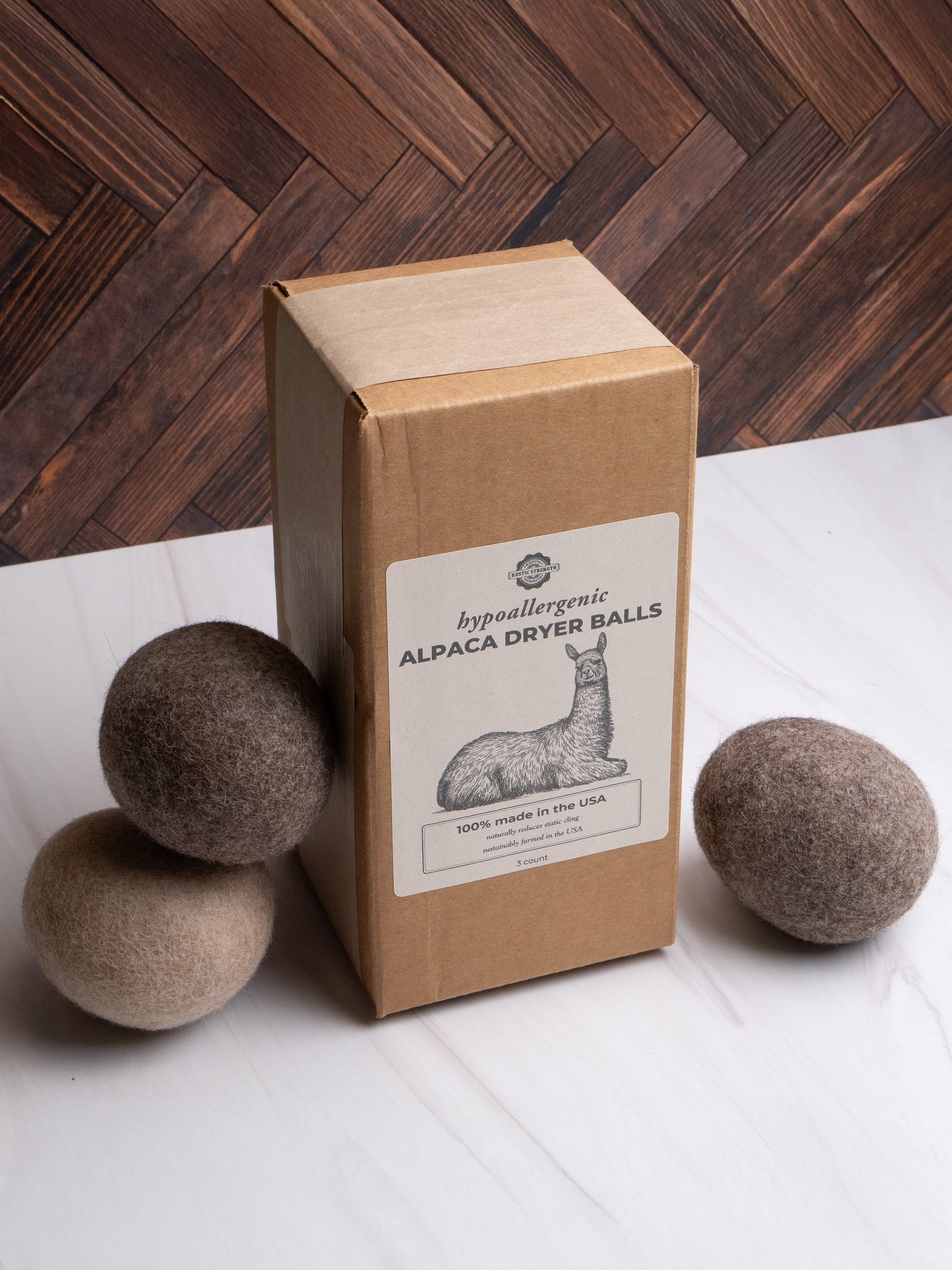 A Rustic Strength Alpaca Dryer Balls (3 pack) box rests on marble, with three brown and gray dryer balls beside it; a wooden herringbone background suggests natural static reduction.