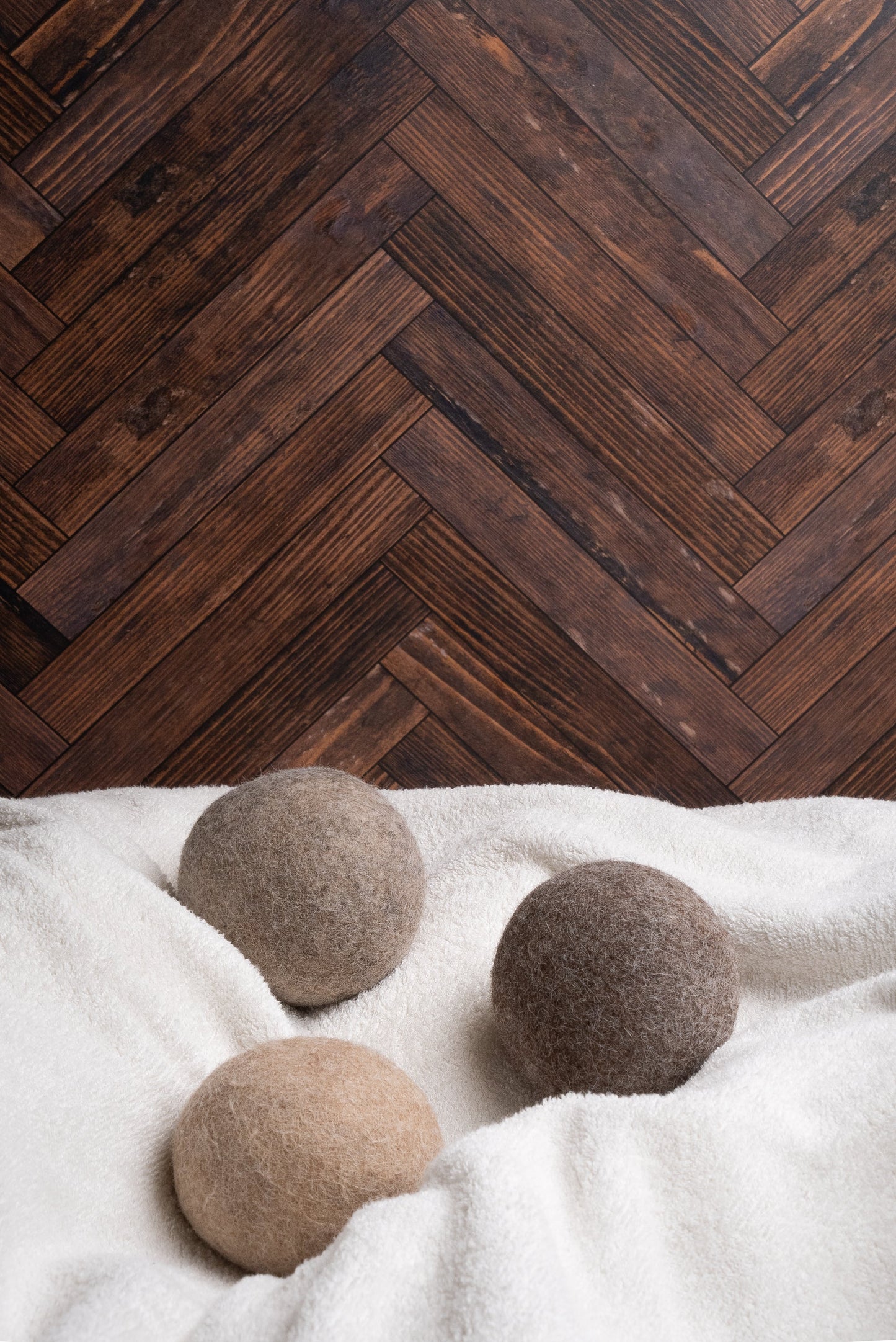Rustic Strength’s Alpaca Dryer Balls (3 pack) in beige, brown, and gray rest on a white towel, providing a natural wool alternative with static reduction. The scene is set against a dark wooden herringbone-patterned floor.