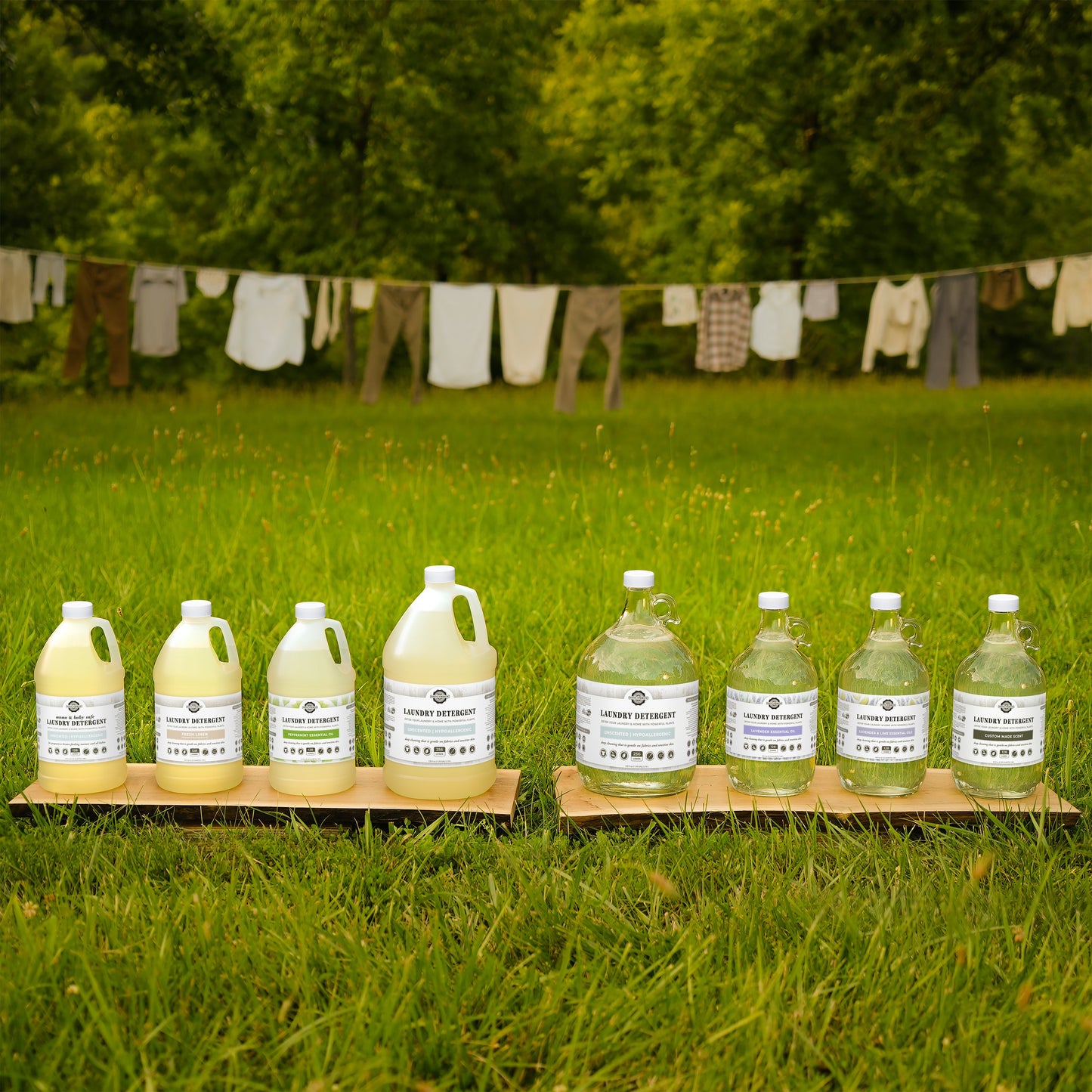 Large bottles of Rustic Strength Powder Laundry Detergent are arranged on wooden boards in a grassy field, with trees and laundry drying on a clothesline in the background.