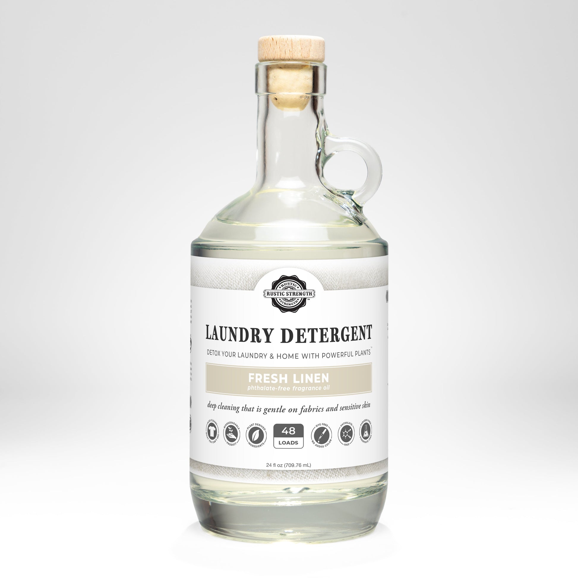 A clear glass bottle with a wooden cork holds Rustic Strength Laundry Detergent | Fresh Linen, 24 fl oz (48 loads). The label highlights plant-based, hypoallergenic cleaning with icons for gentle, biodegradable detergent.